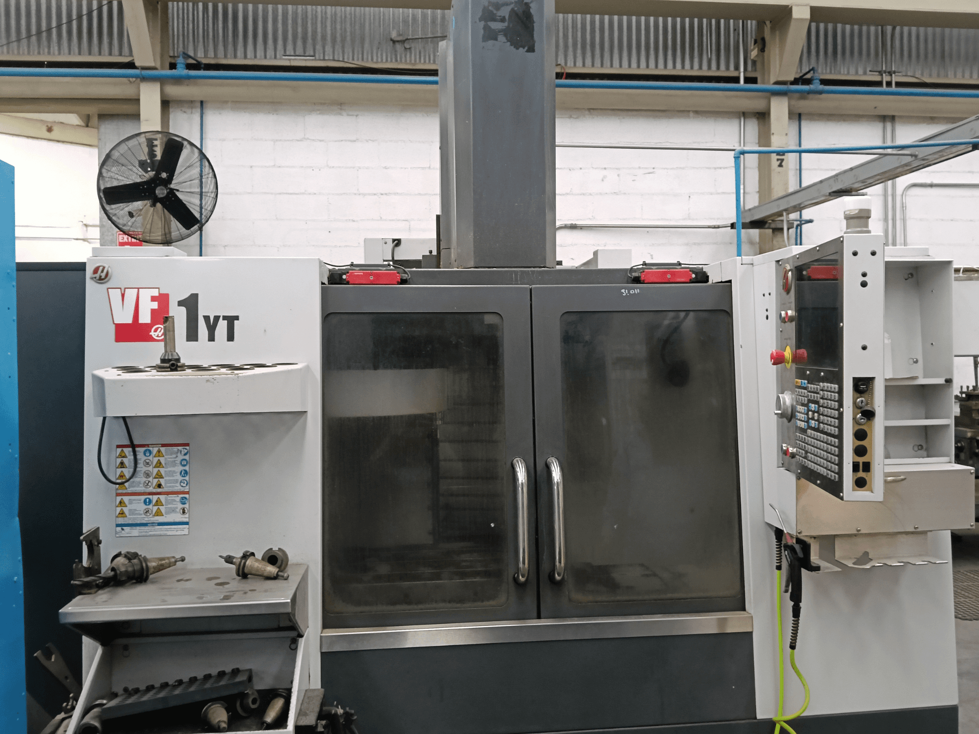 HAAS VF-1YT-1 vertikaliojo apdirbimo centro vaizdas iš priekio su valdymo pultu ir šone esančiais įrankiais.