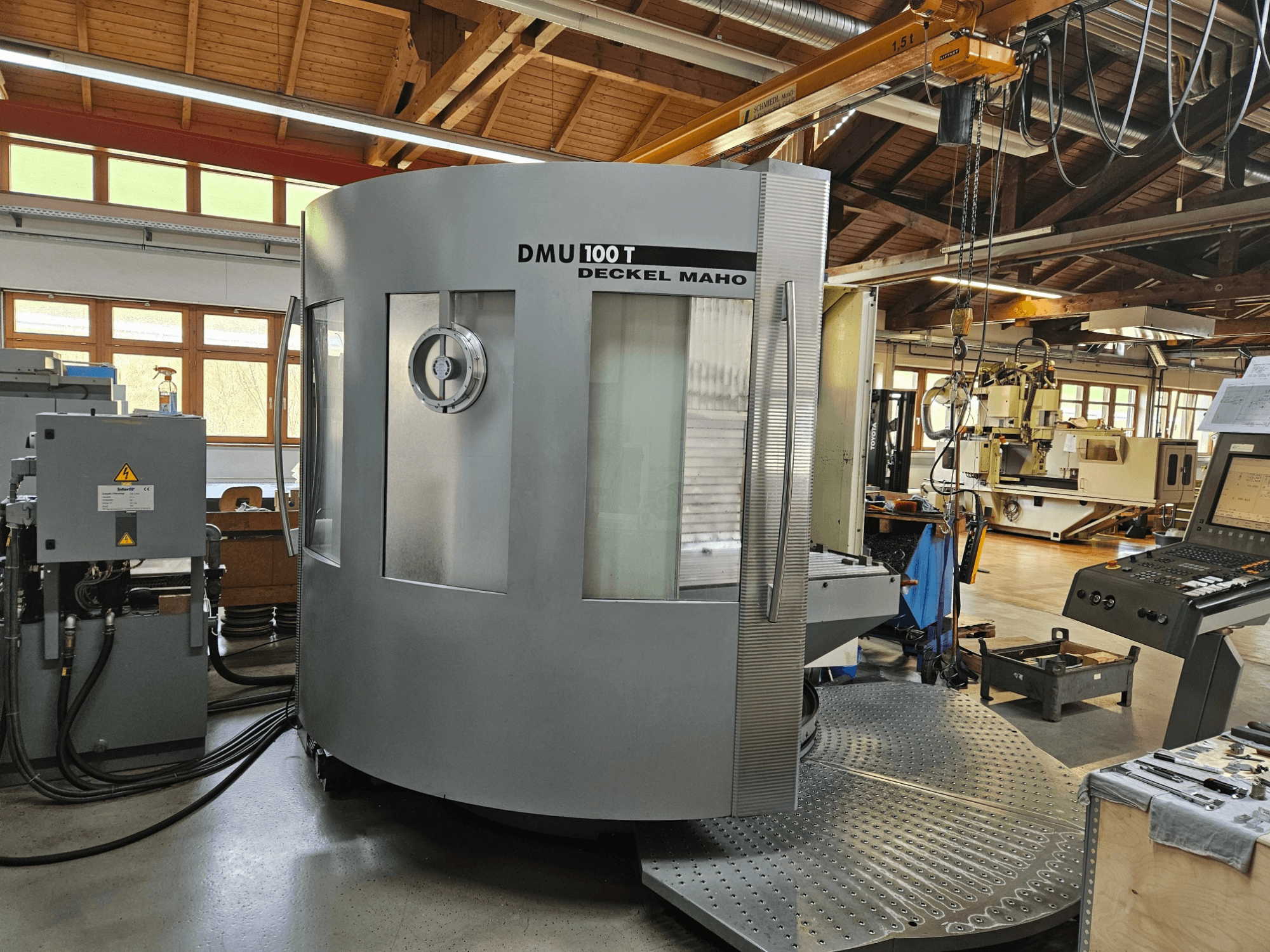 DMU 100 T Deckel Maho CNC staklės, žiūrint iš priekio, su dideliu apvaliu pagrindu, valdymo skydeliu ir stikliniu korpusu.