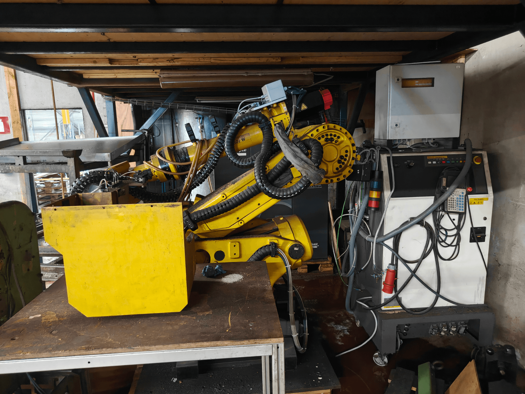 FANUC roboto S-430i W rankos vaizdas iš priekio: geltona, su valdymo pultu, kabeliais ir geltonu komponentu ant darbastalio.