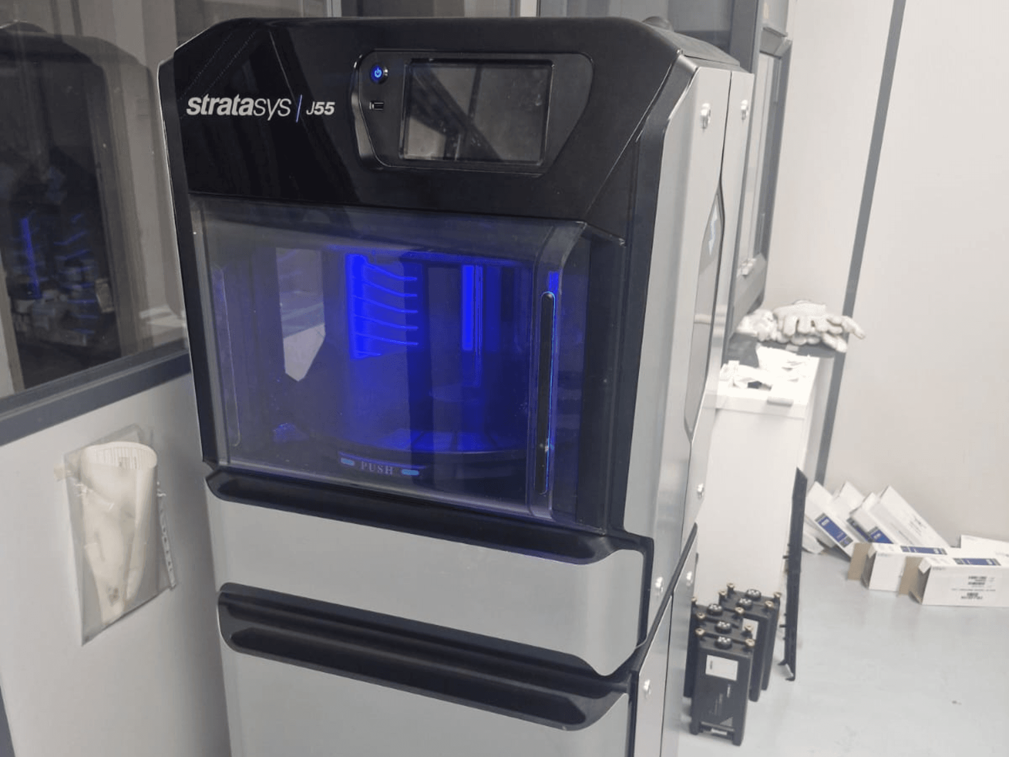 Plastikinio 3D spausdintuvo "Stratasys J55 Prime" šoninis vaizdas, kuriame matomas jo aptakus dizainas ir apšviesta spausdinimo sritis.