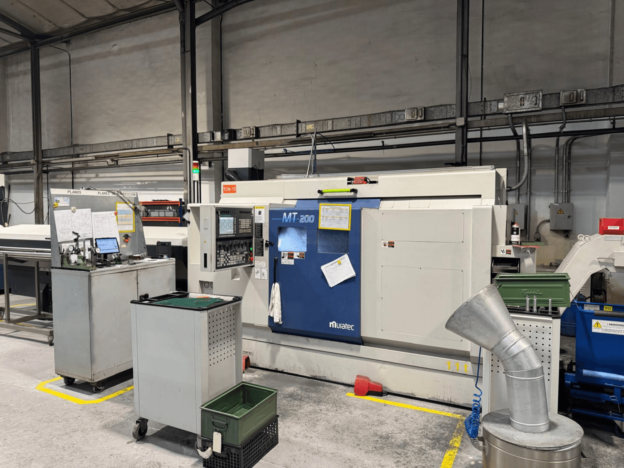 Gamyklinės Muratec MT200 daugiaverpstės tekinimo staklės, žiūrint iš priekio, su valdymo skydeliu ir aplinkine įranga.
