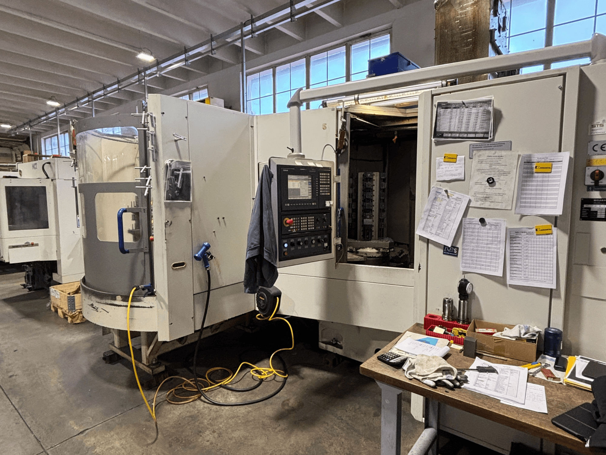 CNC staklės dirbtuvėse, vaizdas iš priekio, šone - valdymo skydelis ir dokumentai; įrankiai ir medžiagos ant stalo.