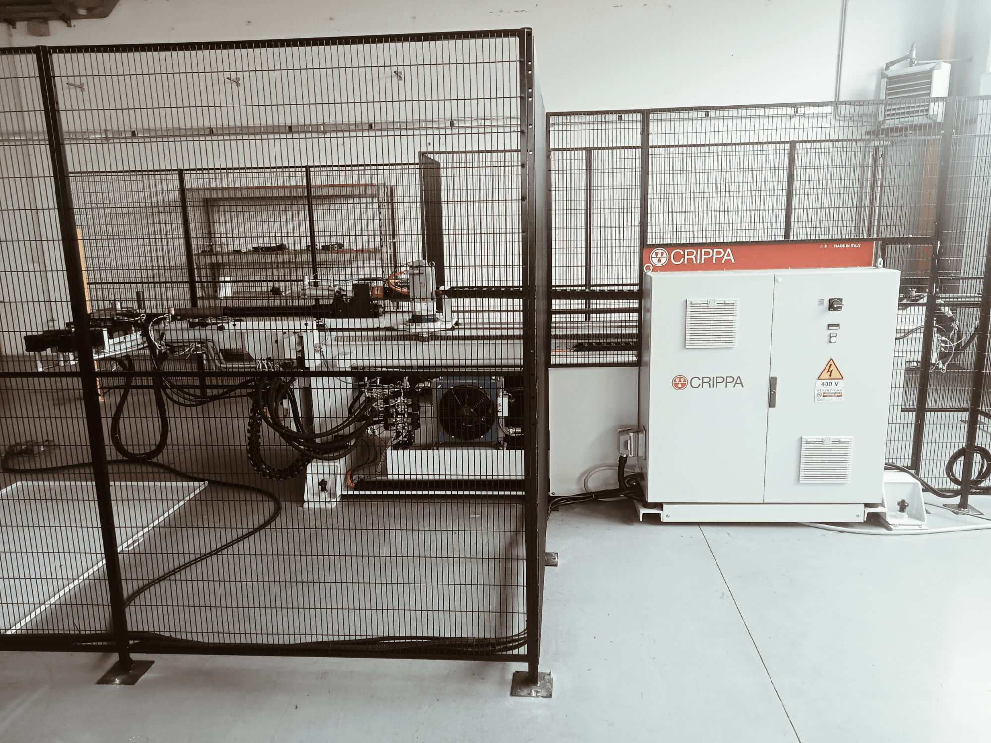 Balta CRIPPA mašina už apsauginio narvo, su matomomis žarnomis ir komponentais, vaizdas iš šono, pramoninė aplinka.