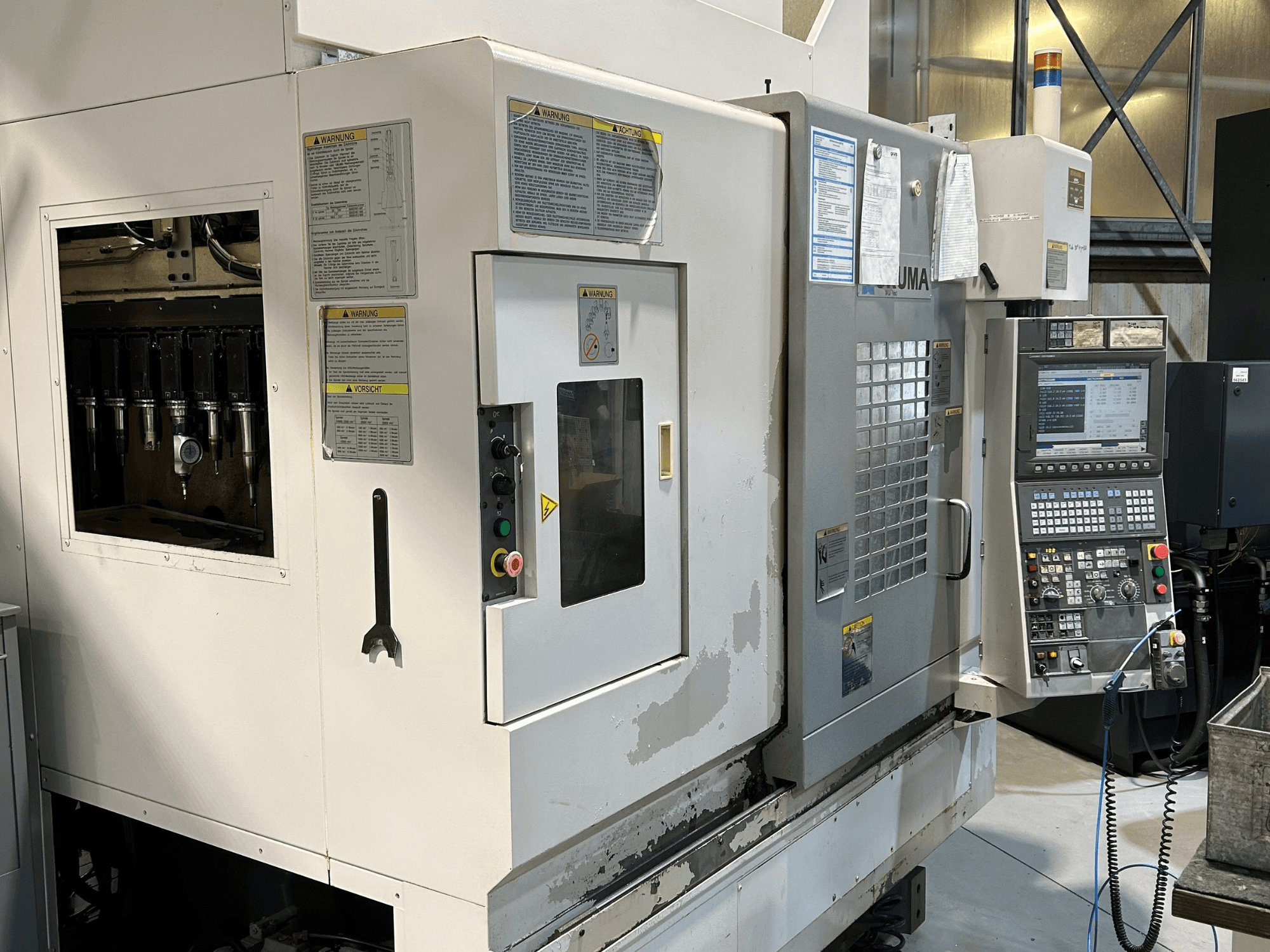 UMA CNC staklių vaizdas iš priekio su matomais įrankių laikikliais, valdymo skydeliu, įspėjamaisiais užrašais ir valdymo mygtukais.