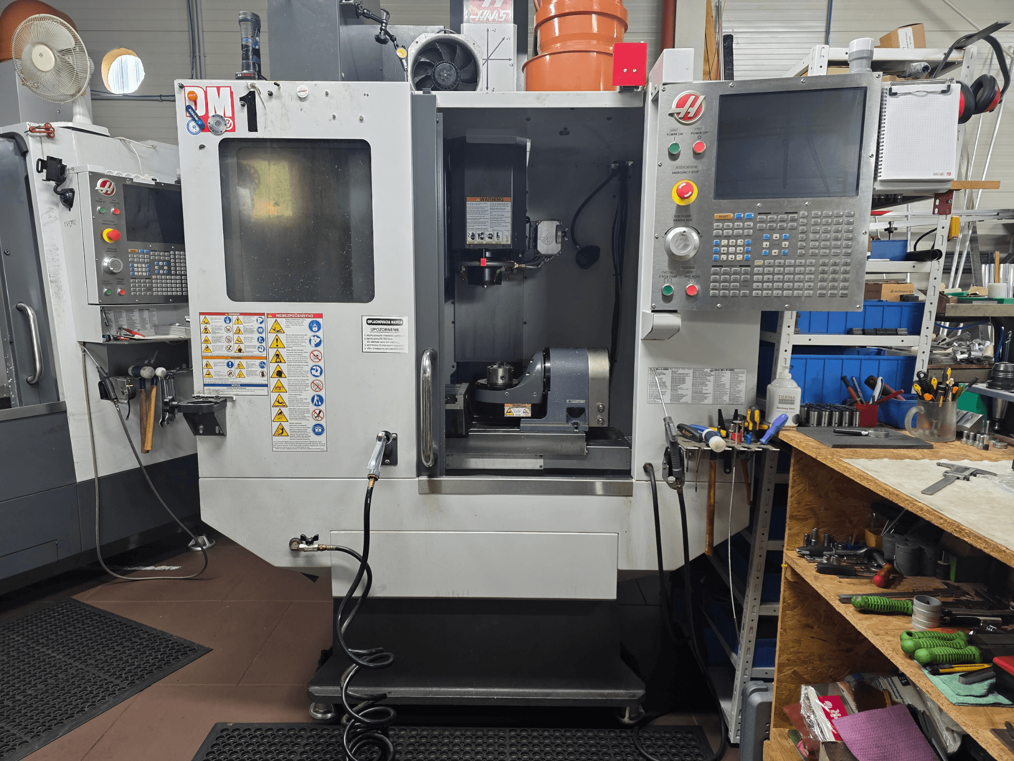 Haas CNC staklių modelis VF-2, vaizdas iš priekio, kuriame matyti valdikliai, suklio sritis, saugos etiketės ir įrankiai ant netoliese esančio darbo stalo.