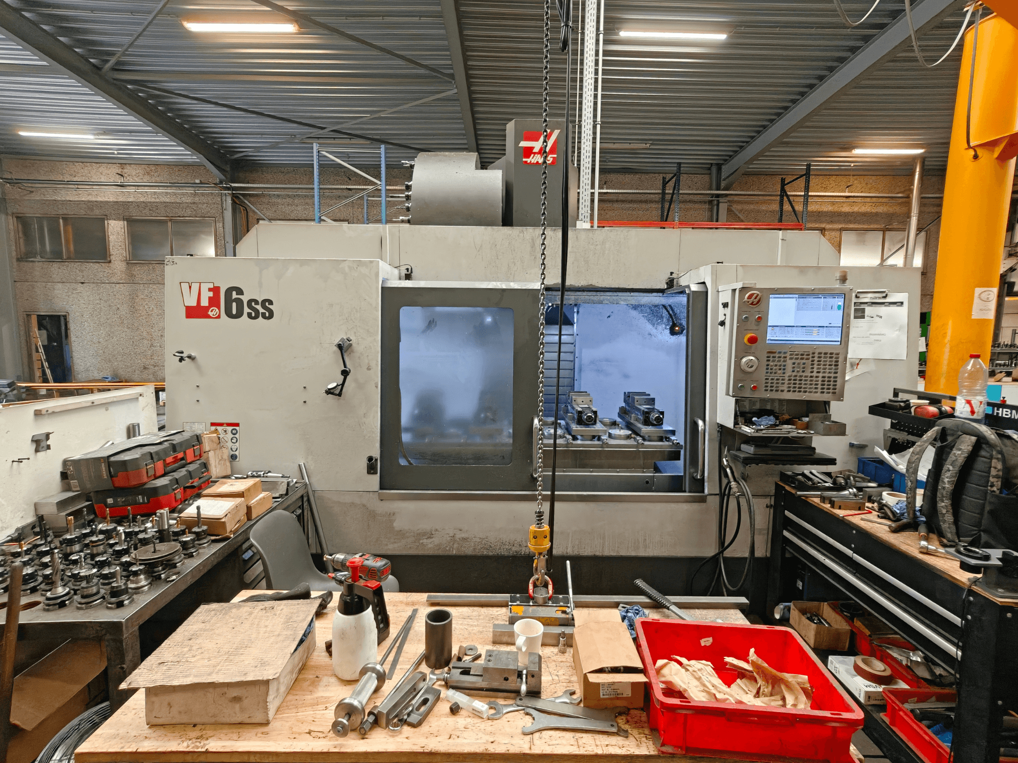 Vertikalusis apdirbimo centras „Haas VF-6SS“ dirbtuvėse, vaizdas iš šono, kuriame matomas valdymo pultas ir darbo vieta, užstatyta įrankiais.
