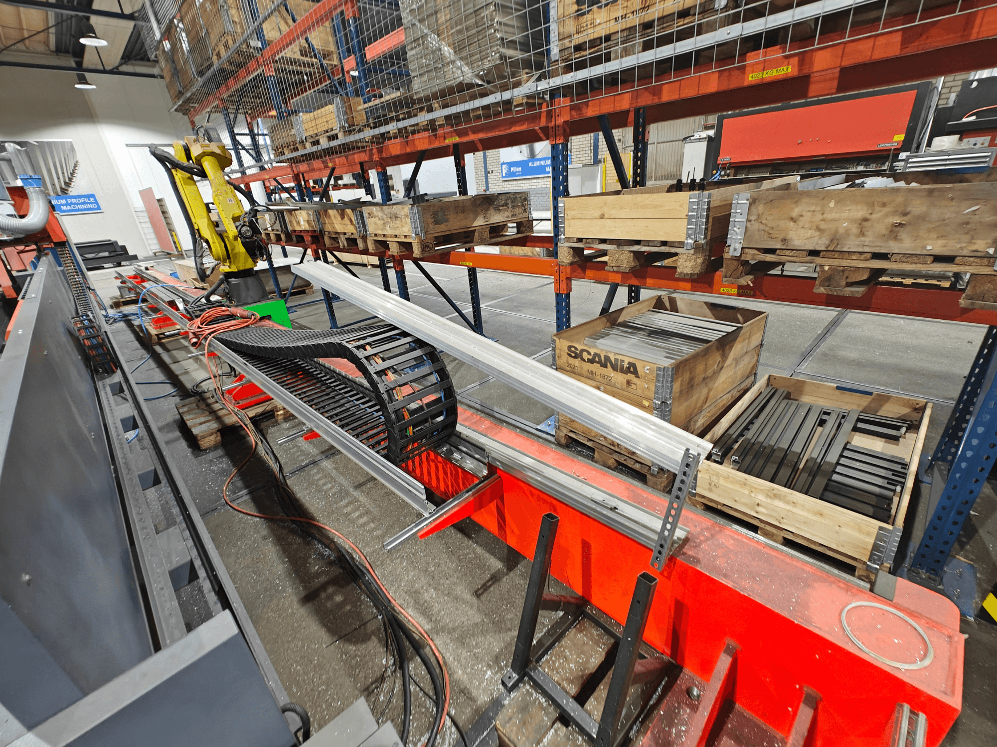 Roboto ranka ant darbo vietos, matomi oranžiniai ir juodi kabeliai, su "Scania" dėžėmis ir aliuminio profiliais gamykloje.