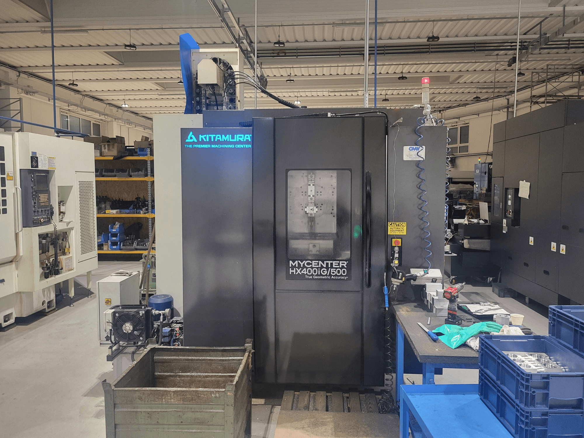 Kitamura Mycenter HX400iG/500 CNC staklės dirbtuvėse, vaizdas iš priekio, šalia yra valdymo įtaisai, įrankių laikymo vieta ir medžiagos.