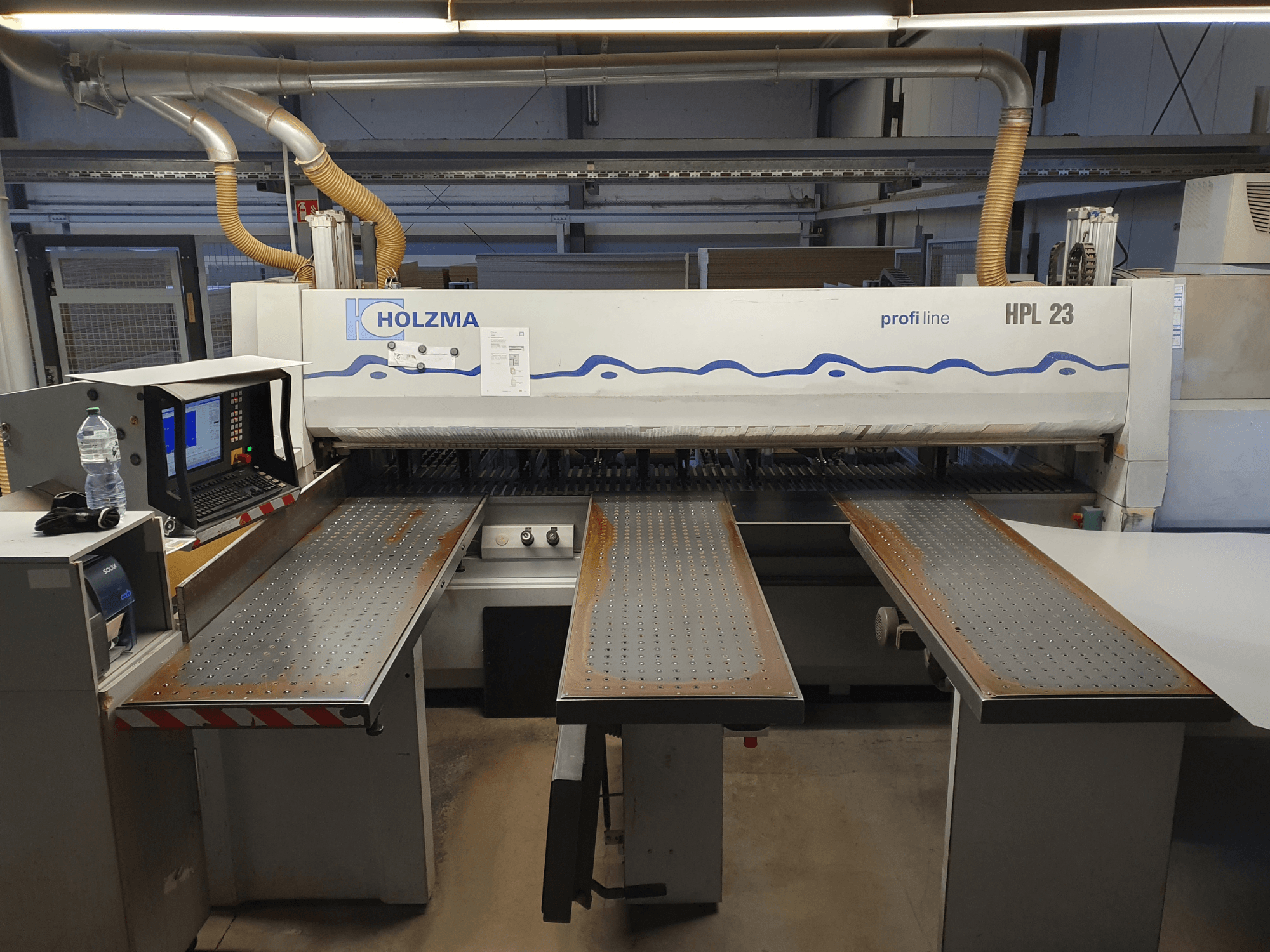 HOLZMA HPL 23 pjaustymo staklės, parodytos iš priekio, su trimis perforuotais stalais, kompiuterio terminalu ir vandens buteliu šalia.