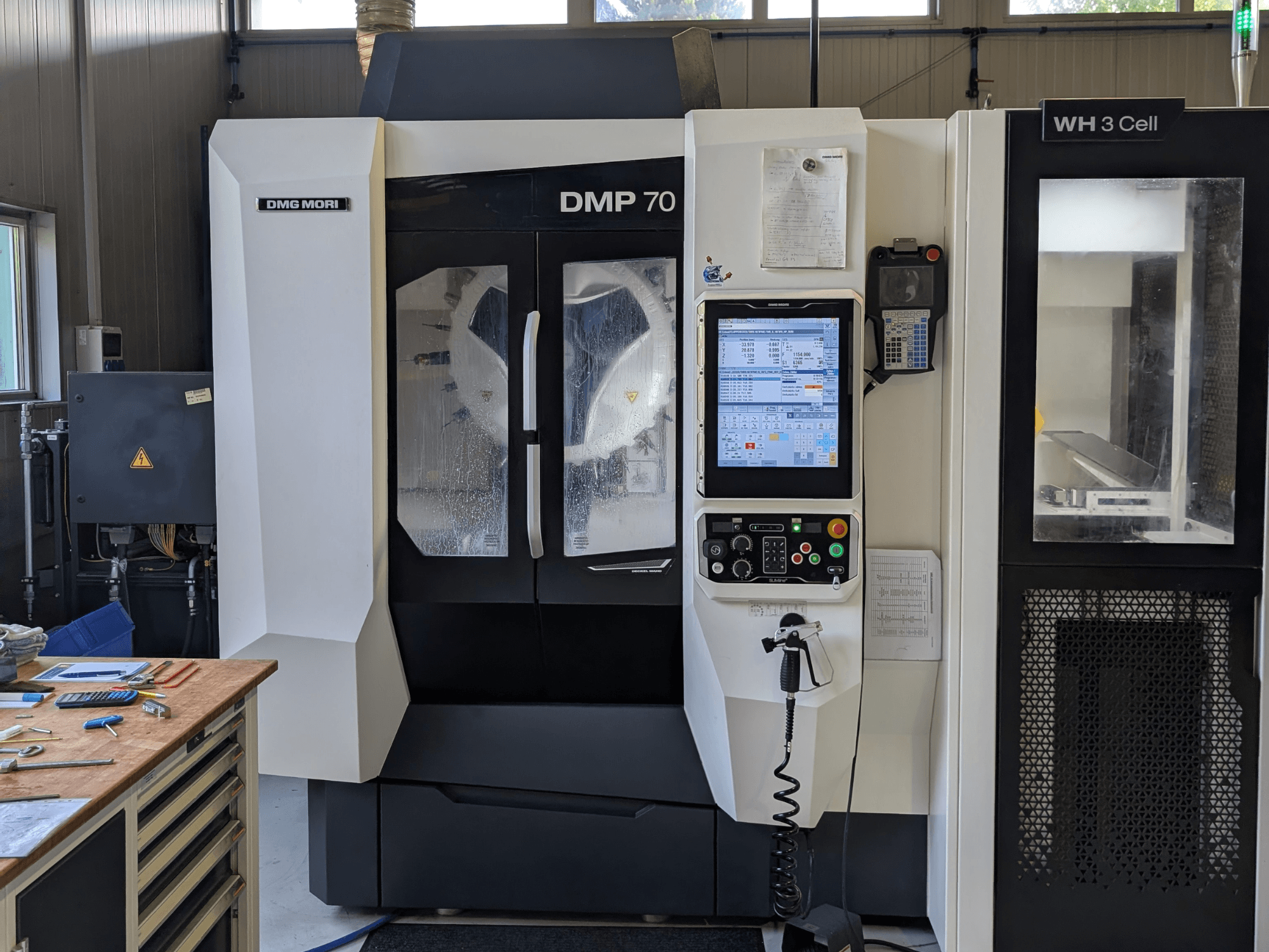 Vertikalus apdirbimo centras „DMG MORI DMP 70“, vaizdas iš priekio su valdymo pultu ir darbo ekranu, apsuptas dirbtuvės įrankiais.