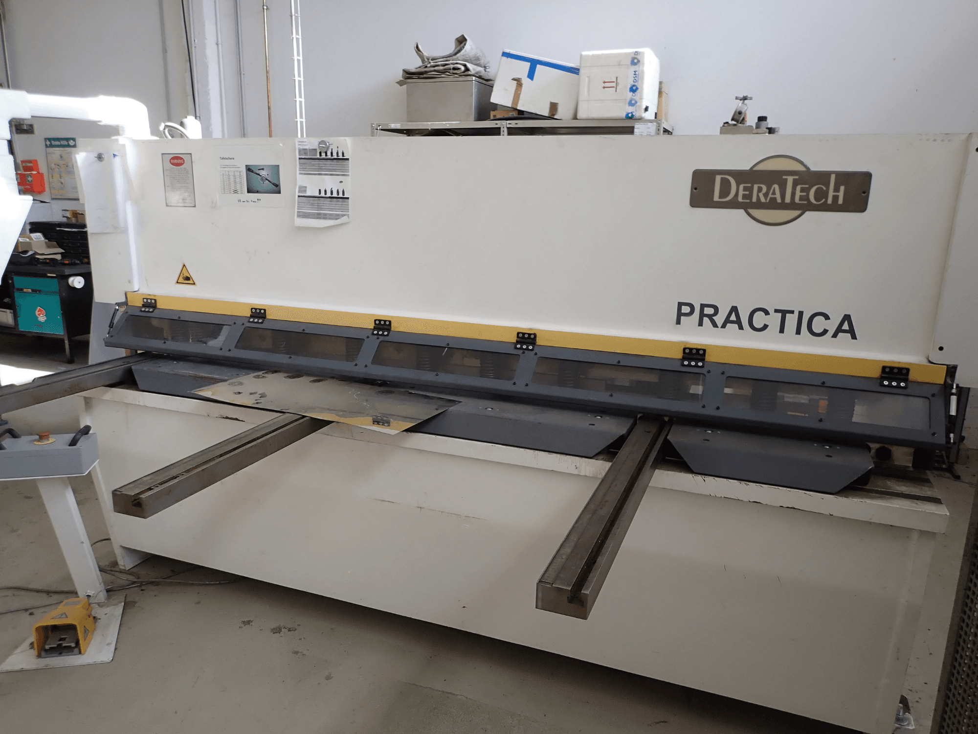 DERATECH Practica CNC kirpimo staklės, vaizdas iš priekio, pjovimo peilis ir metalo lakštai ant darbinio paviršiaus.