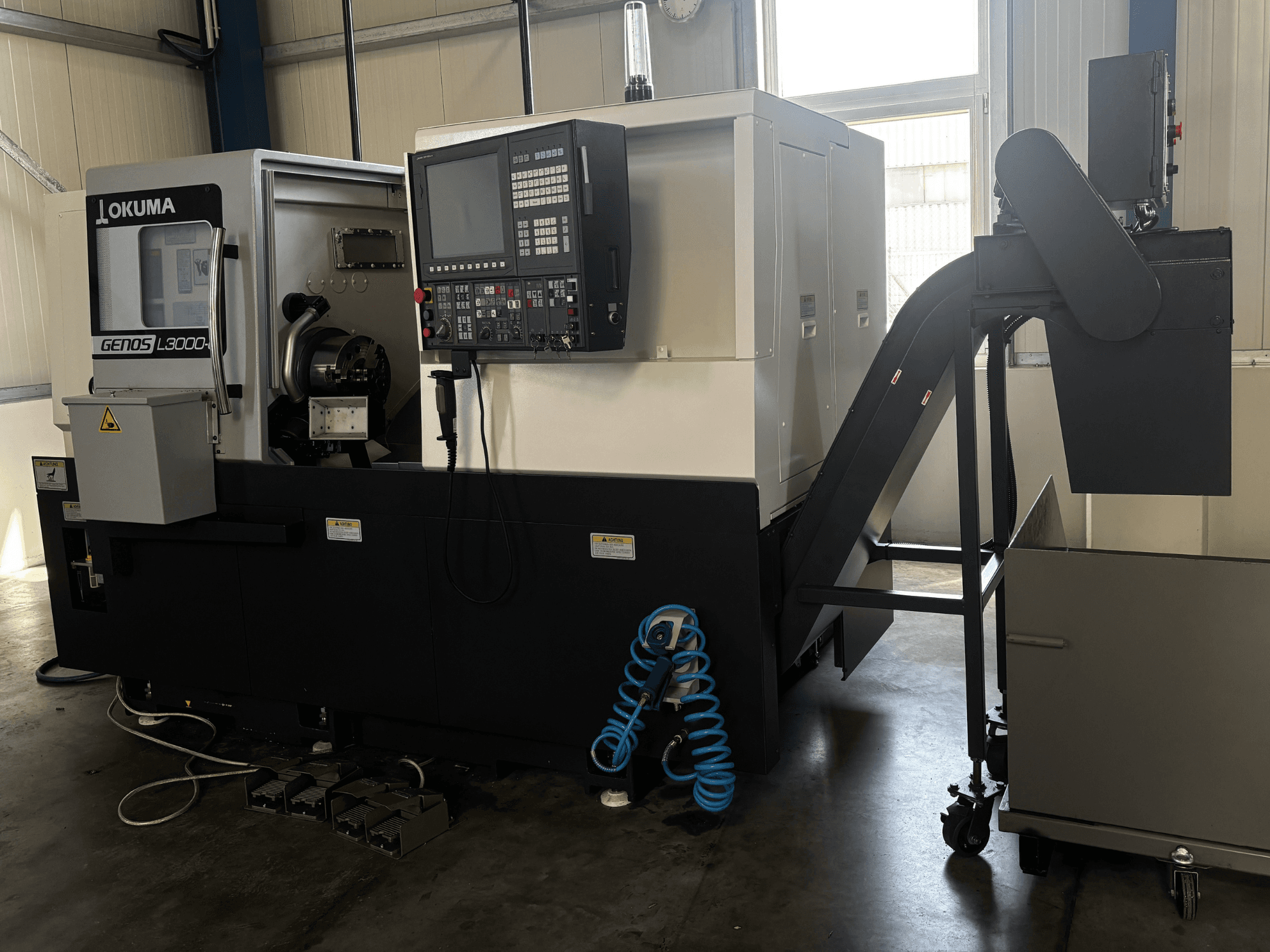 „Okuma GENOS L3000-e“ horizontalusis tekinimo staklės, vaizdas iš priekio, kuriame matomas valdymo pultas, konvejeris ir suktuvo zona.
