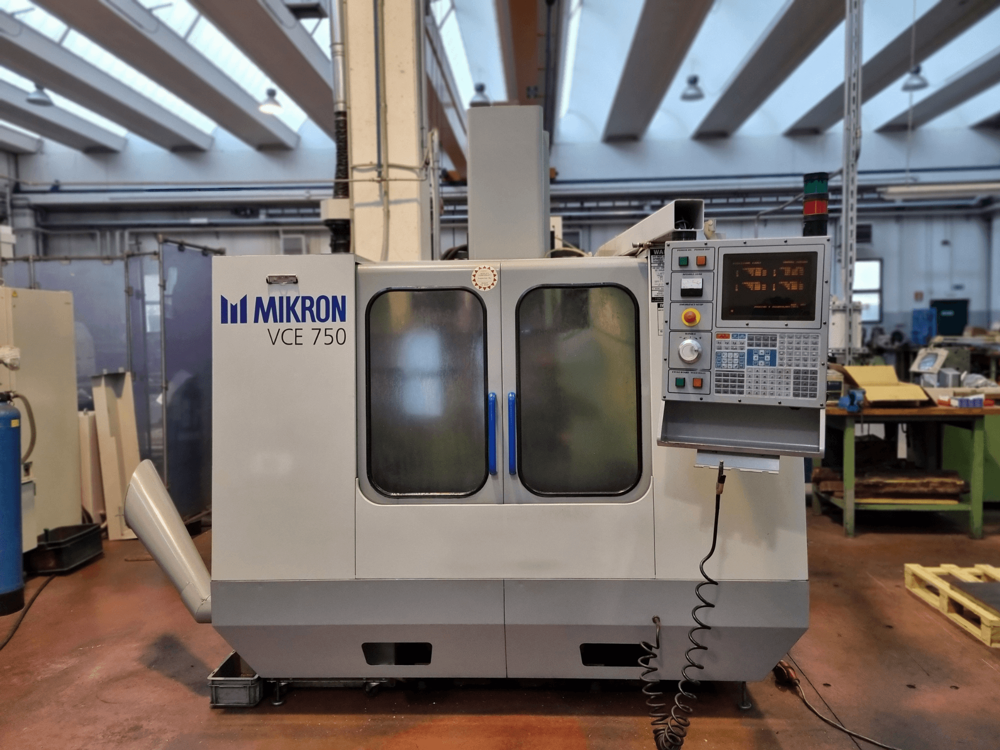 Baltos "Mikron VCE 750 CNC" staklės dirbtuvėse, vaizdas iš šono, su skaitmeniniu valdymo pultu ir didelėmis stiklinėmis durimis.