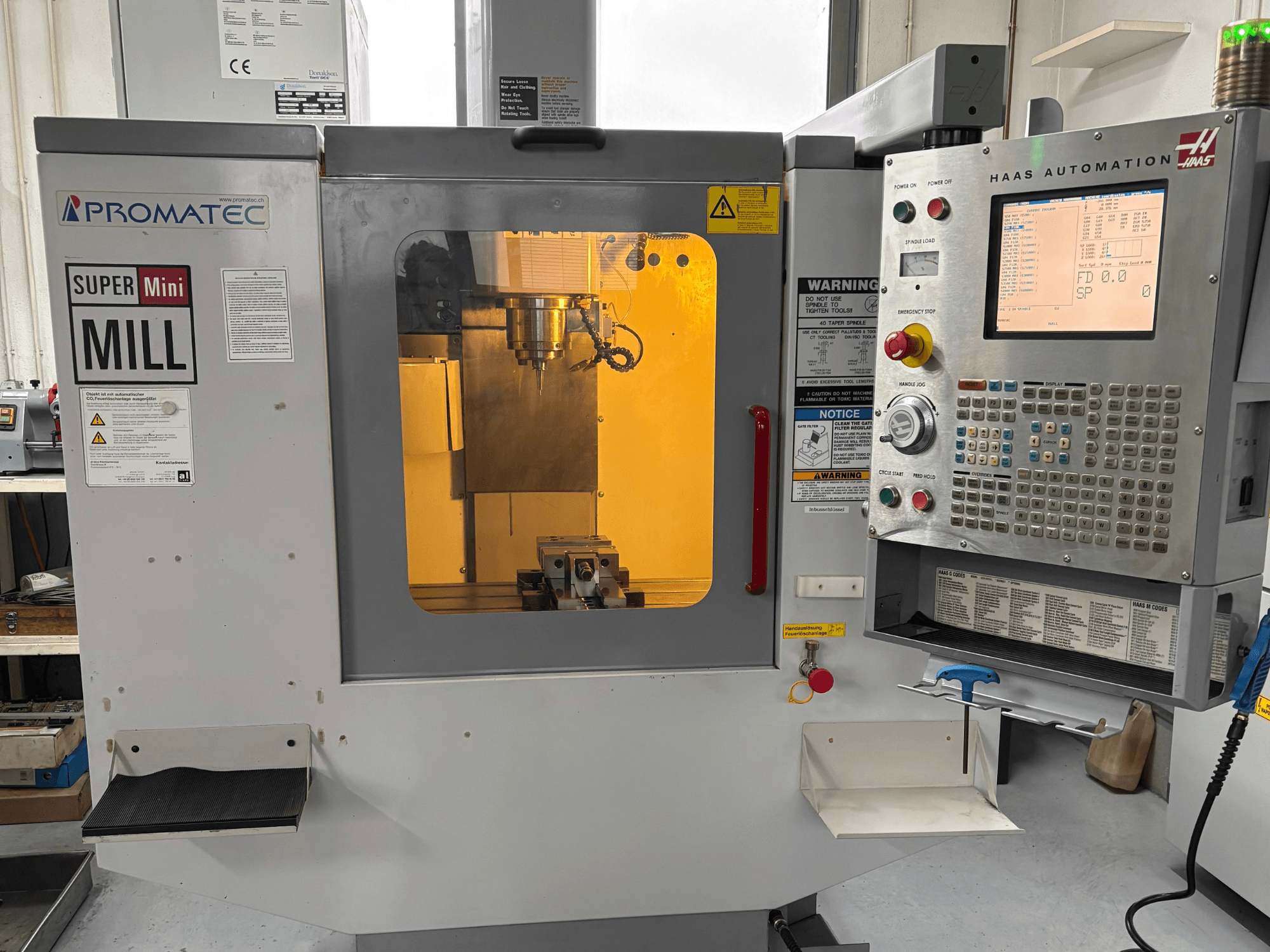 "Promatec Super Mini Mill" frezavimo staklės iš priekio su "Haas Automation" valdymo pultu ir viduje matomu pjovimo įrankiu.