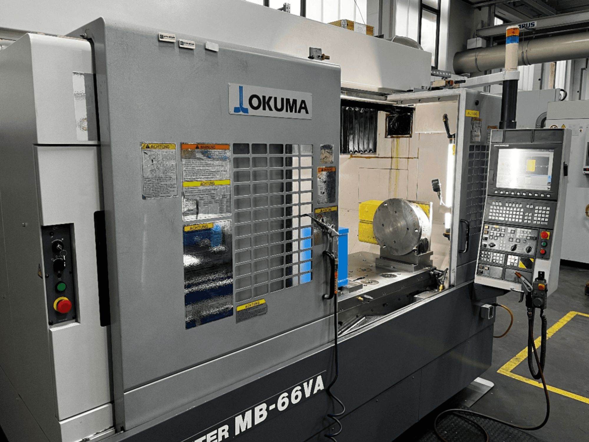 "Okuma MB66-VA" vertikaliojo apdirbimo centro priekinis vaizdas, kuriame matomas valdymo skydelis, ruošinys ir staklių vidus su saugos etiketėmis.
