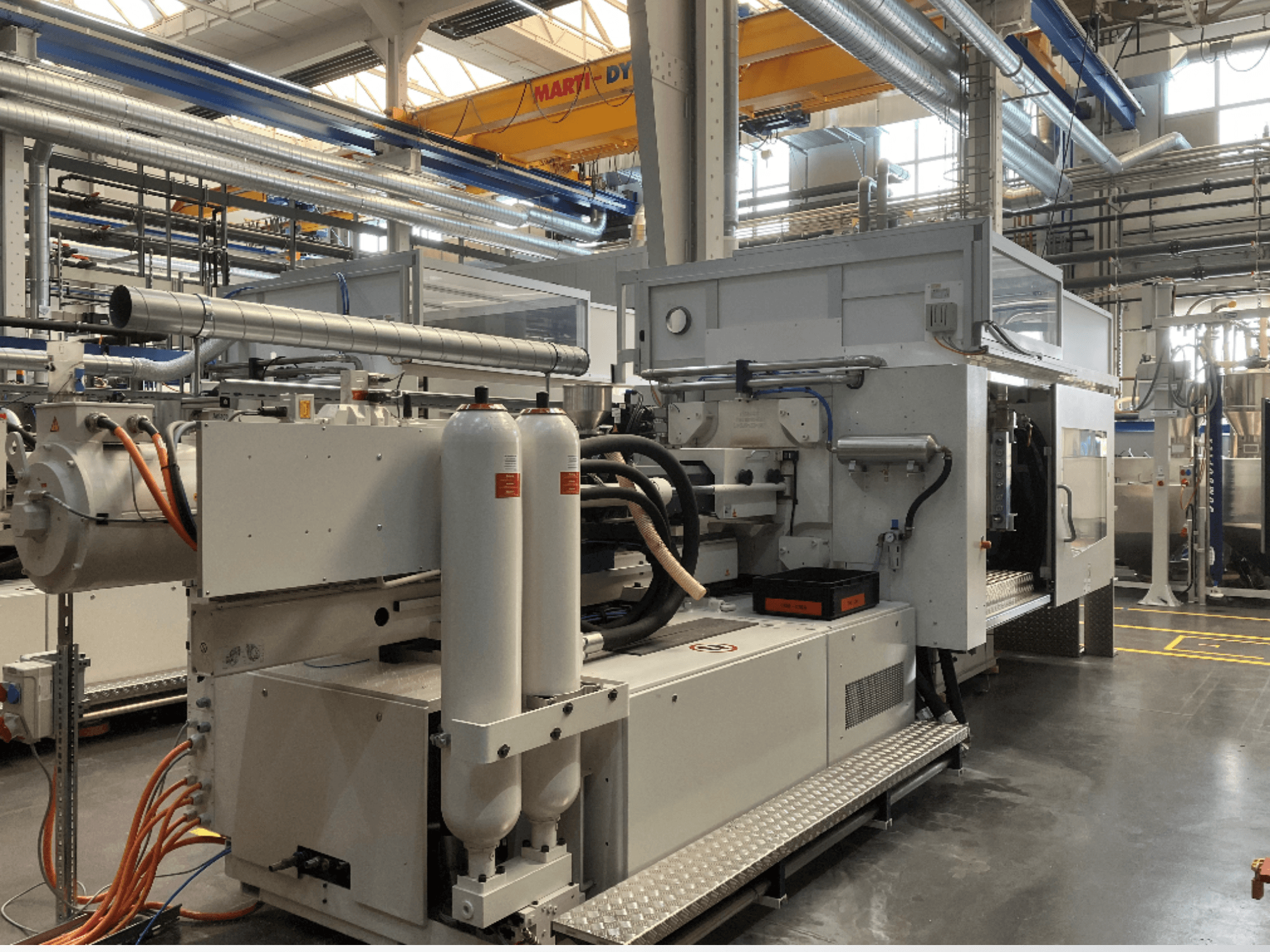 Pramoninė mašina, parodyta iš šono, su keliomis žarnomis, dviem vertikaliomis talpyklomis ir valdymo pultu; prekės ženklo ir modelio nematyti.