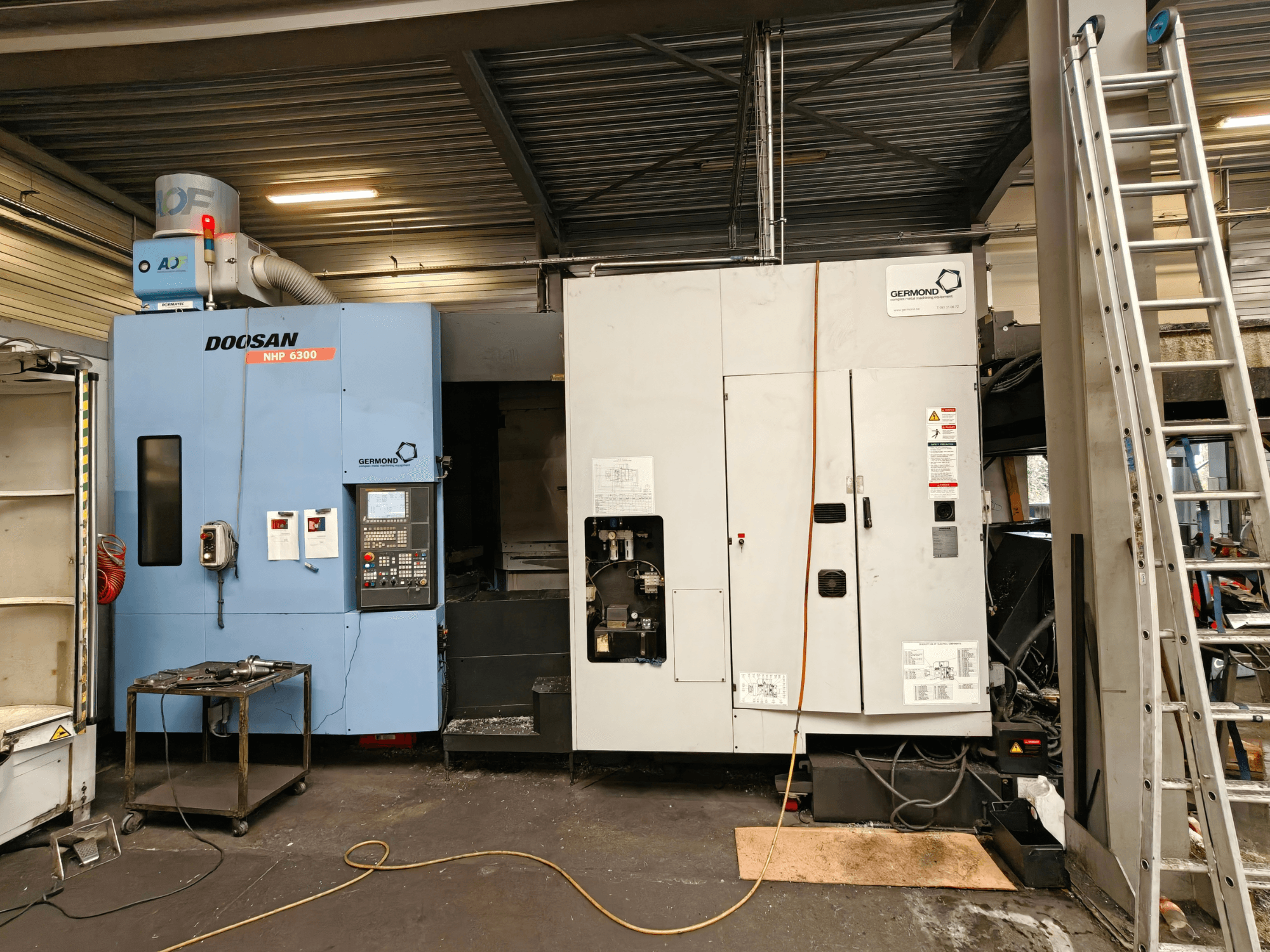 „DOOSAN NHP6300“ horizontalaus apdirbimo centro vaizdas iš priekio, kuriame matomas valdymo pultas ir šalia esantys stakliai dirbtuvės aplinkoje.