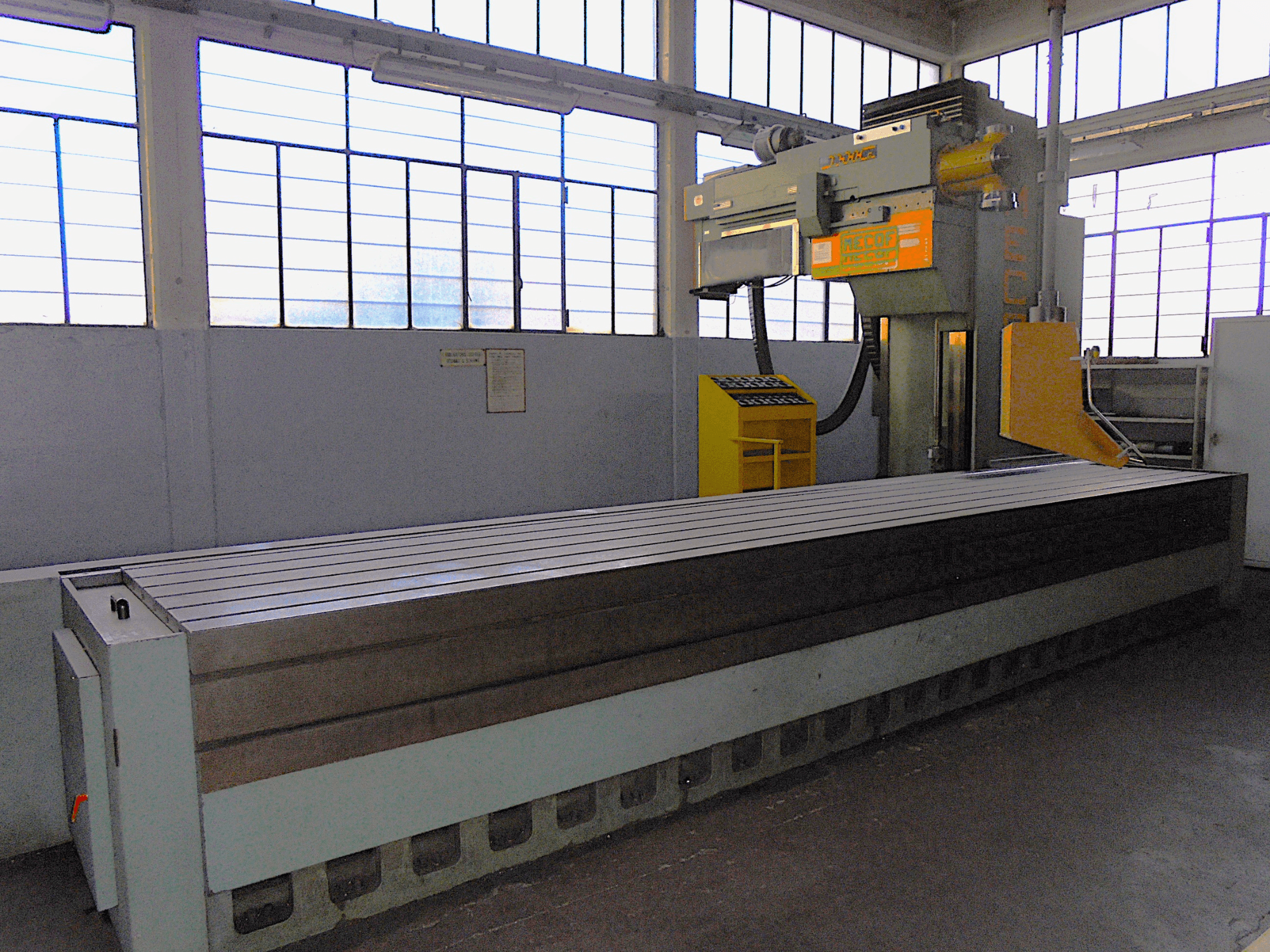 Nežinomos markės vertikalios frezavimo staklės gamykloje, vaizdas iš šono, didelis stalas su grioveliais ir valdymo pultas.
