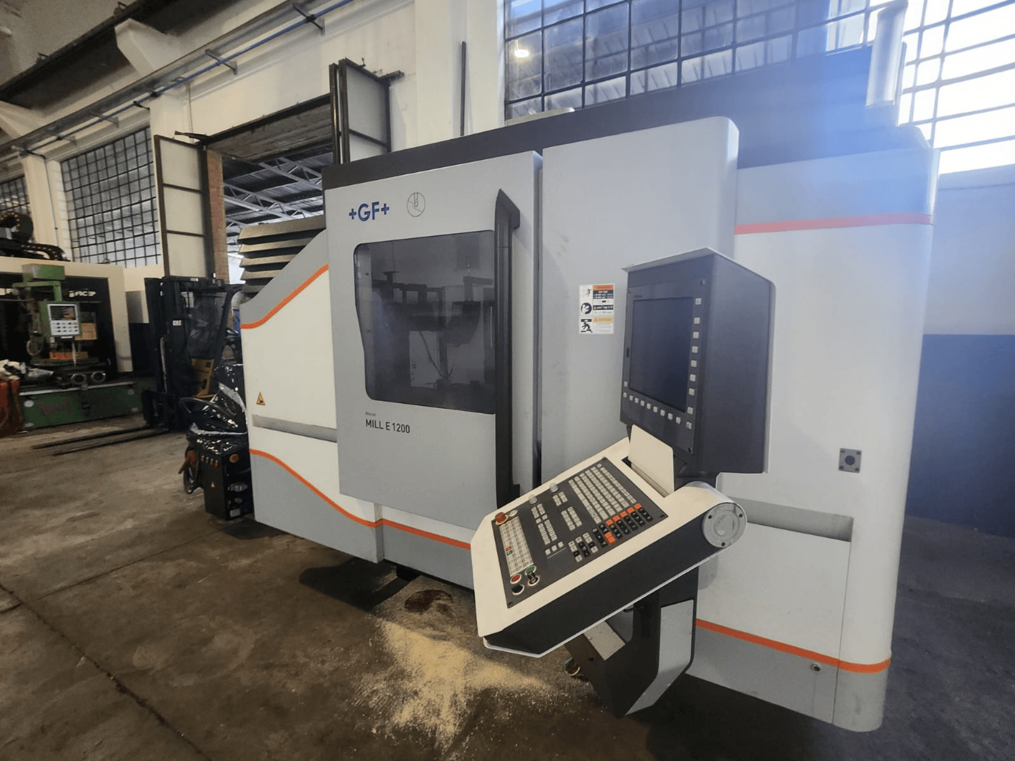 GF MILL E1200, frezavimo staklių vaizdas iš priekio su valdymo skydeliu, ekranėliu ir saugos etiketėmis, dirbtuvių aplinka.