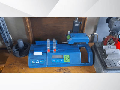 Mėlynas "Hommel T 1000" spausdintuvas ir T 1 010 testeris su raudonais mygtukais, žiūrint iš priekio ant darbo stalo su metaliniais blokais.