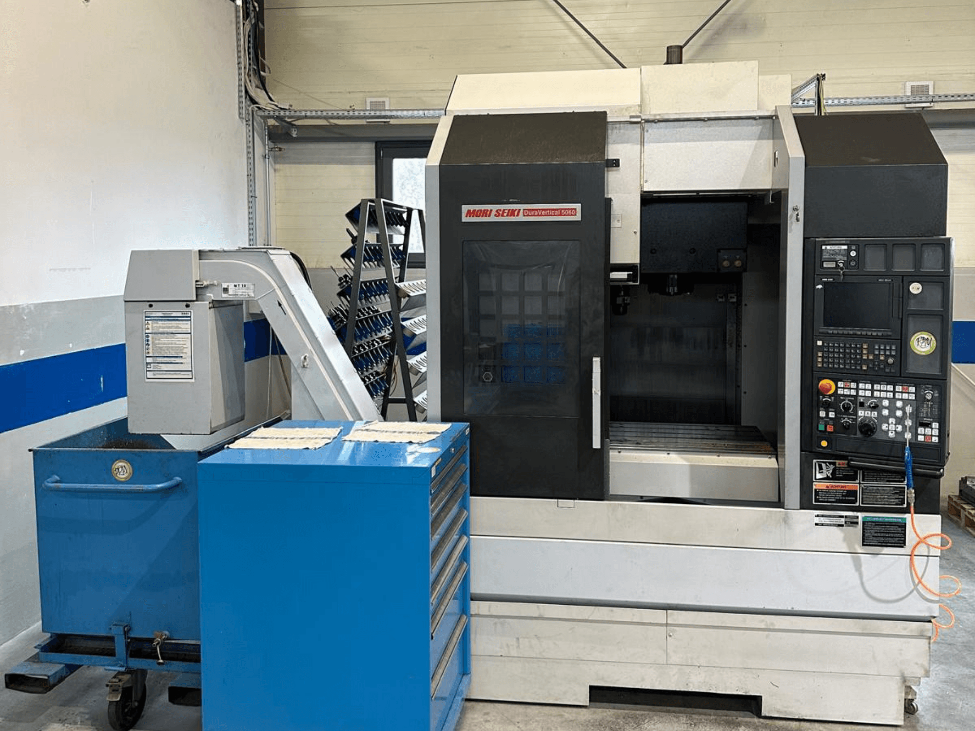 MORI SEIKI DURA VERTICAL 5060 – vertikalaus apdirbimo centro vaizdas iš priekio su valdymo pultu ir šalia esančia įrankių saugykla.