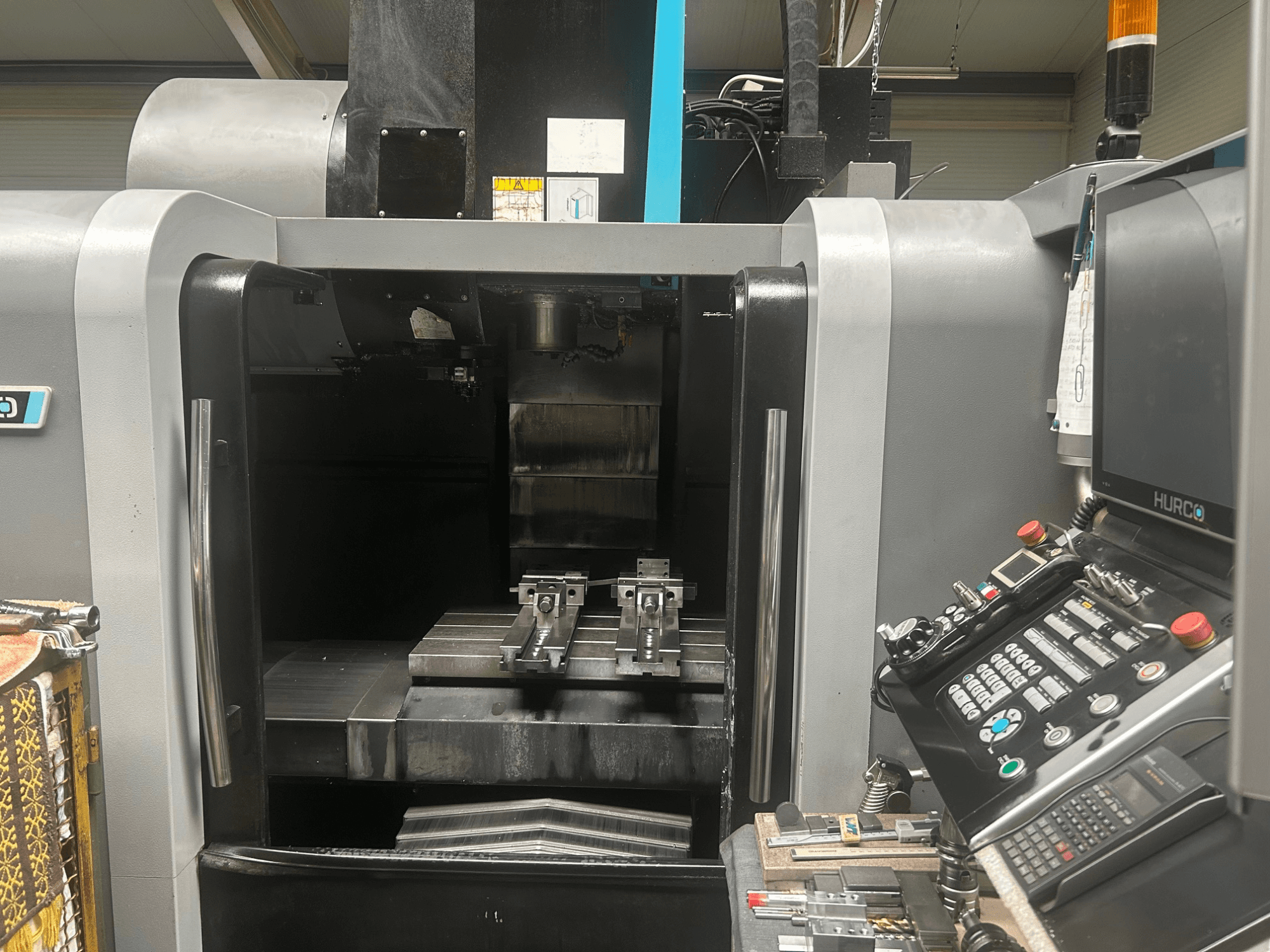CNC apdirbimo centro "Hurco" priekinis vaizdas, kuriame matomas įrankių laikiklis, metalinis ruošinys ir valdymo skydelis su mygtukais bei ekranu.