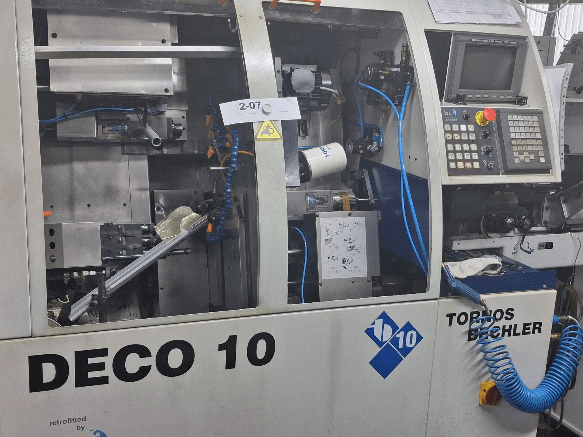 Staklių TORNOS Deco 10a šveicariško tipo tekinimo staklių vaizdas iš arti, matomos vidinio mechanizmo ir valdymo skydelio detalės.