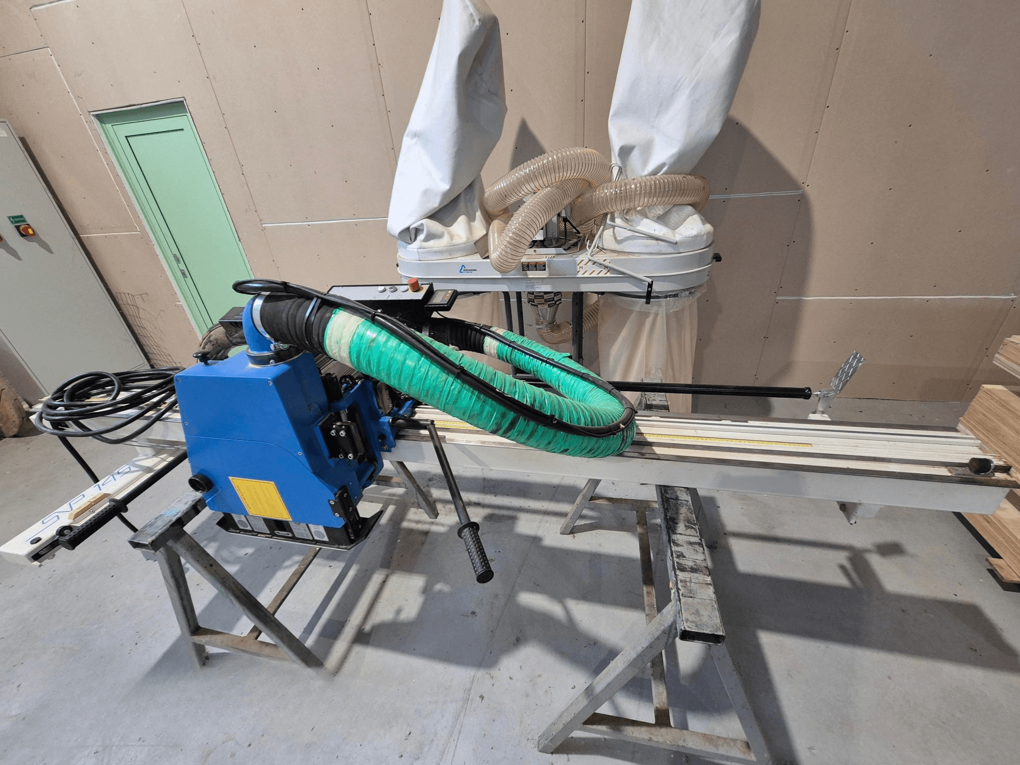 Mėlynas SVP-175 horizontalusis plokščių pjūklas su žalia dulkių žarna ir atrama, žiūrint iš šono dirbtuvėse.
