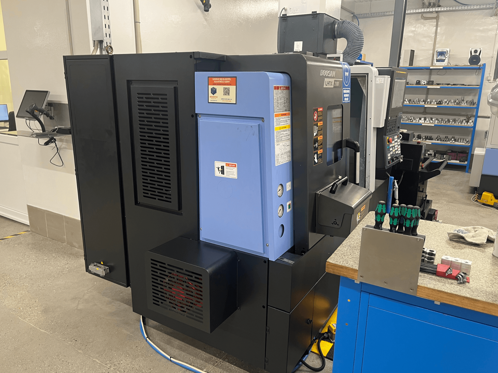 Staklės "Doosan Lynx 2100", šoninis vaizdas, kuriame matyti valdymo skydelis, ventiliacijos sistema ir įrankiai ant darbo stalo.