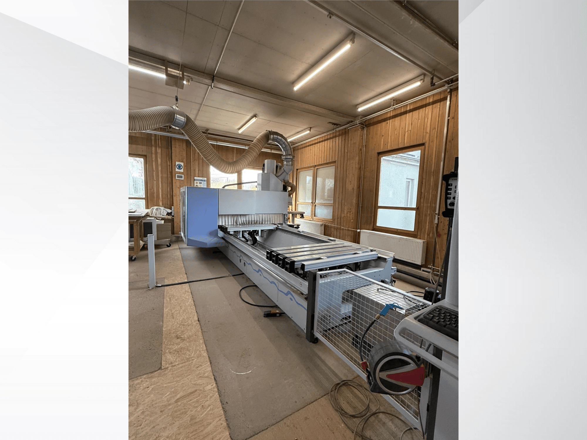 CNC frezavimo staklės dirbtuvėse, šoninis vaizdas, didelis mėlynas korpusas, pritvirtintas juostinis transporteris ir matomas valdymo skydelis.