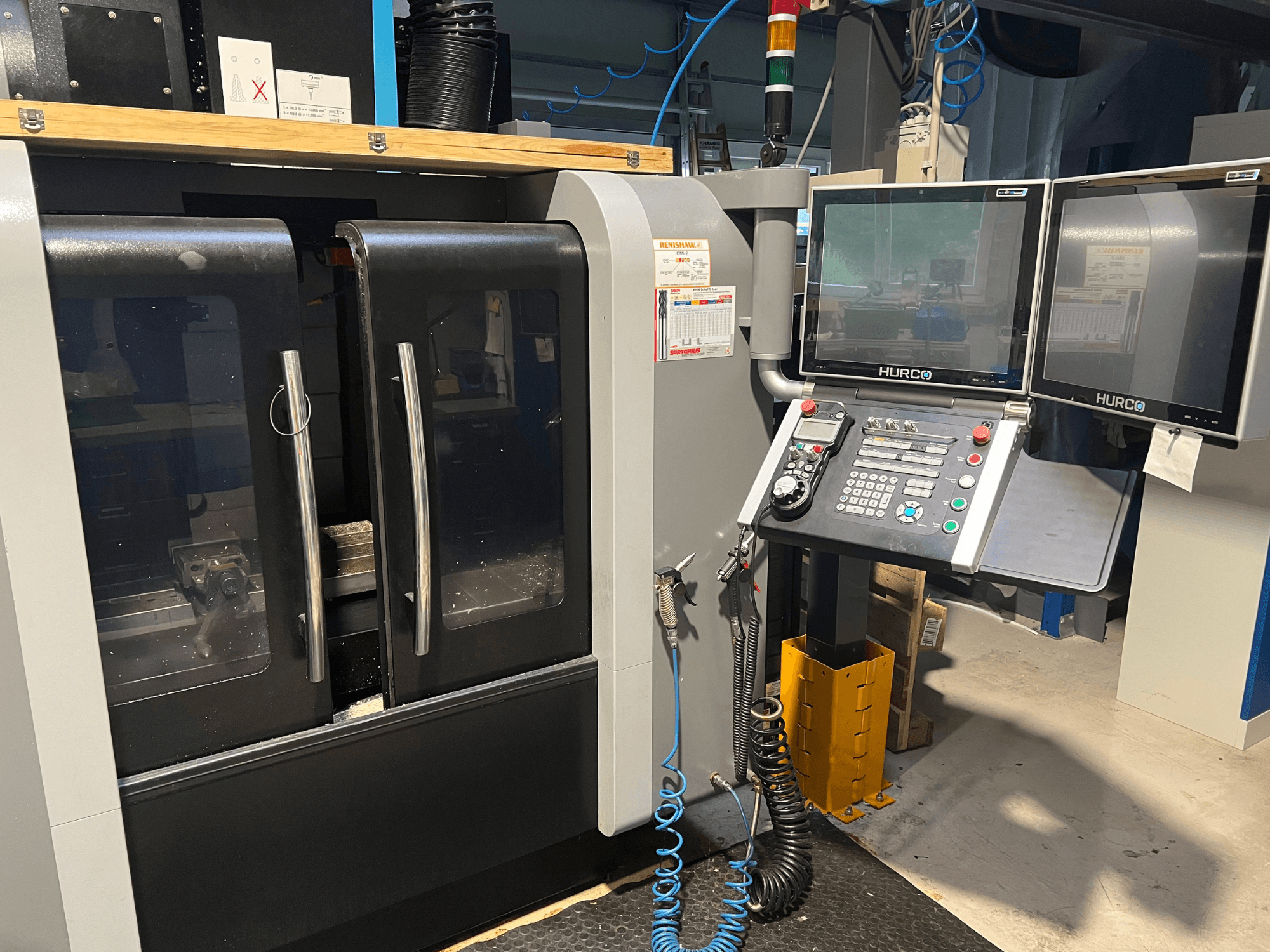Hurco CNC staklių vaizdas iš priekio su dviem monitoriais ir valdymo skydeliu. Skaidrios durys ir viduje matoma apdirbimo zona.