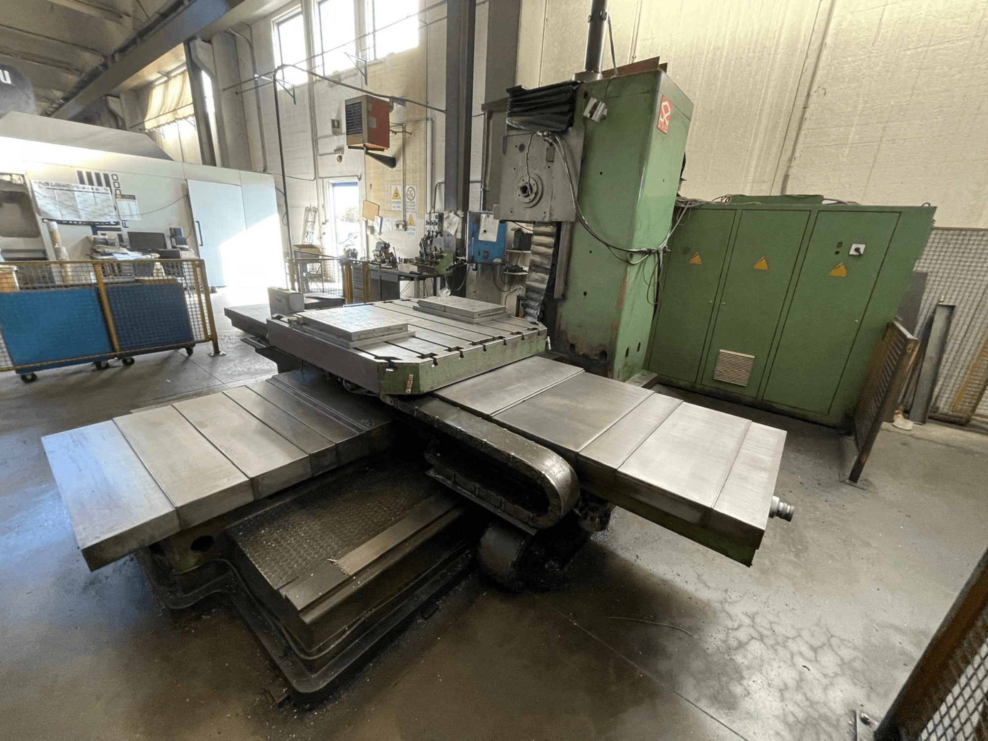 Didelės pramoninės frezavimo staklės žalios spalvos korpusu, vaizdas iš šono, su keliais plokščiais paviršiais ir sandėliavimo spintomis fone.