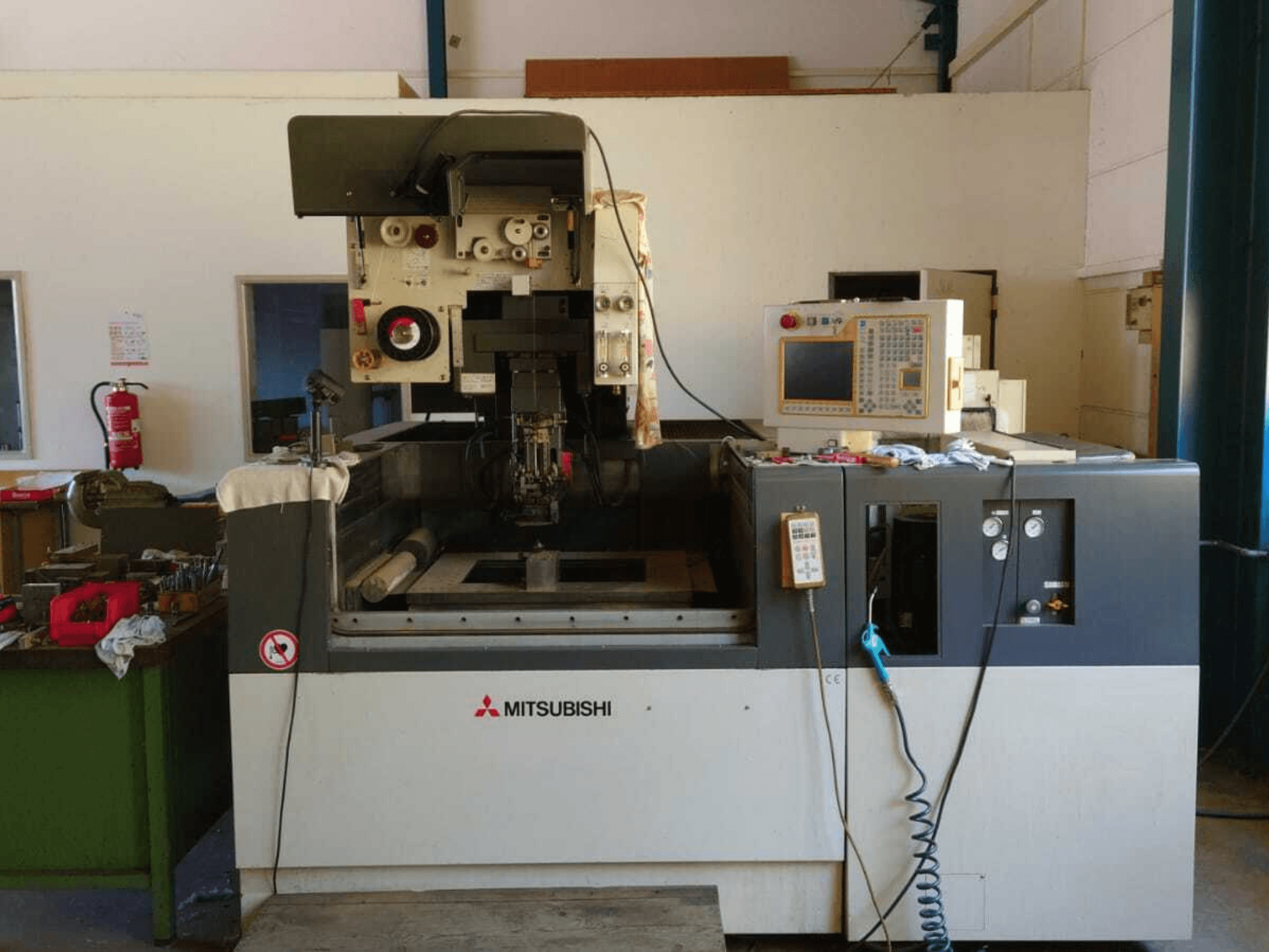 „Mitsubishi FA20“ vielinės elektroerozijos staklių vaizdas iš priekio, kuriame matomas valdymo pultas, darbo zona ir saugos ženklai.