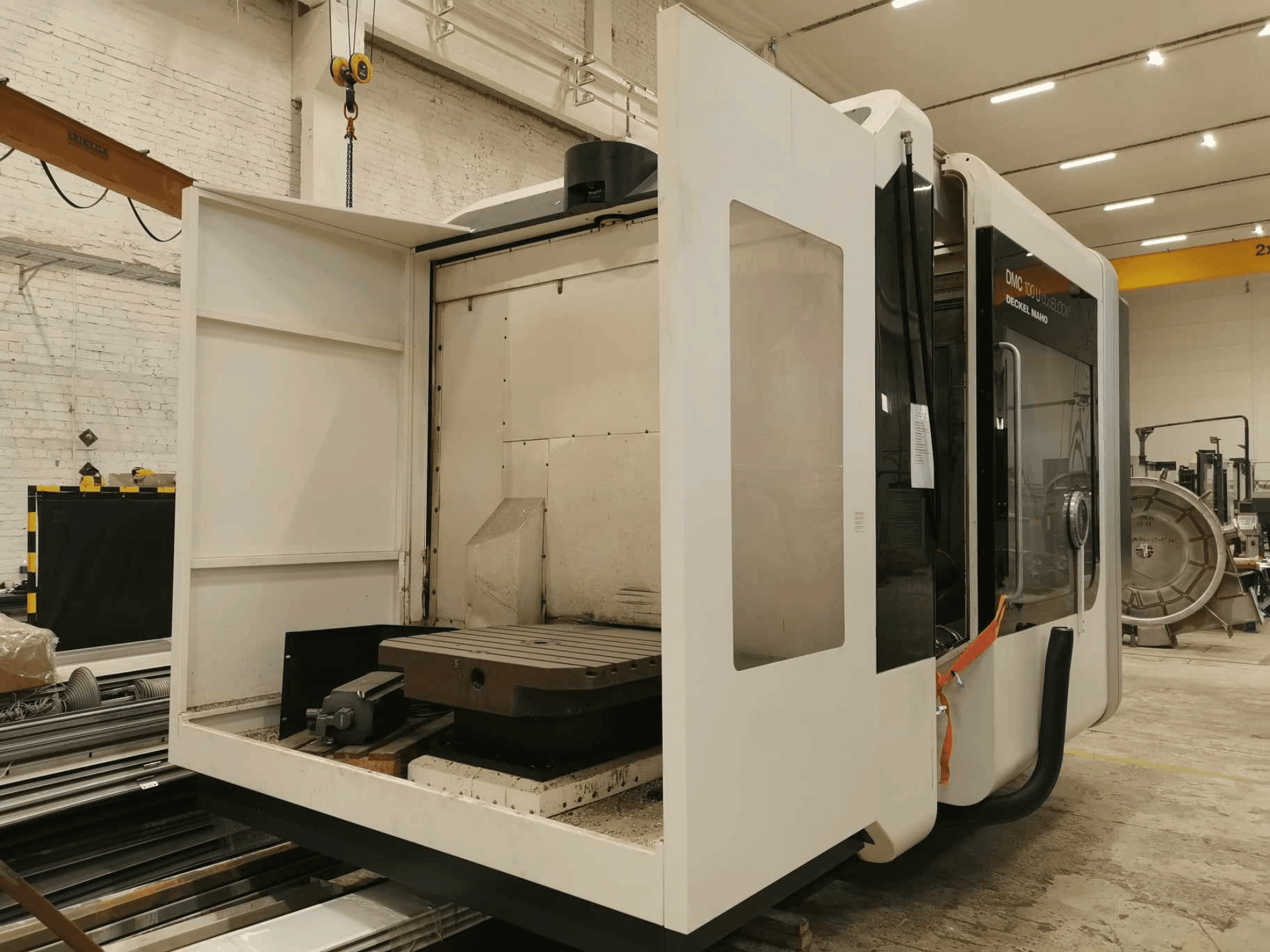 DMC 100U linijinės CNC staklės, šoninis vaizdas su atidarytomis įėjimo durelėmis, matomas darbo stalas ir vidiniai komponentai.