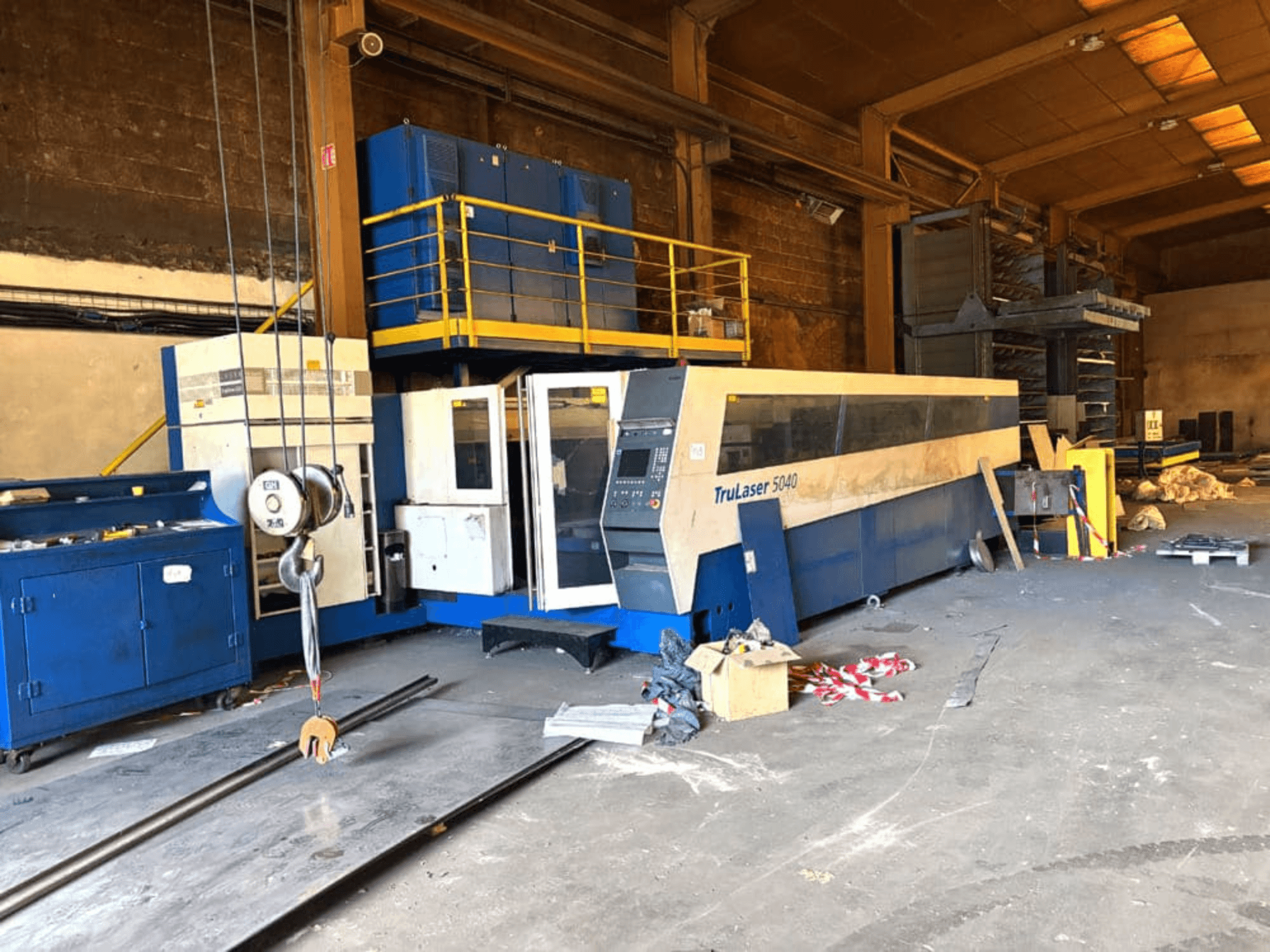 „TRUMPF“ CO₂ lazerio pjovimo staklės, modelis „TRULASER 5040“, nufotografuotos dirbtuvėse, vaizdas iš priekio kampu, fone matosi daiktų gausa.