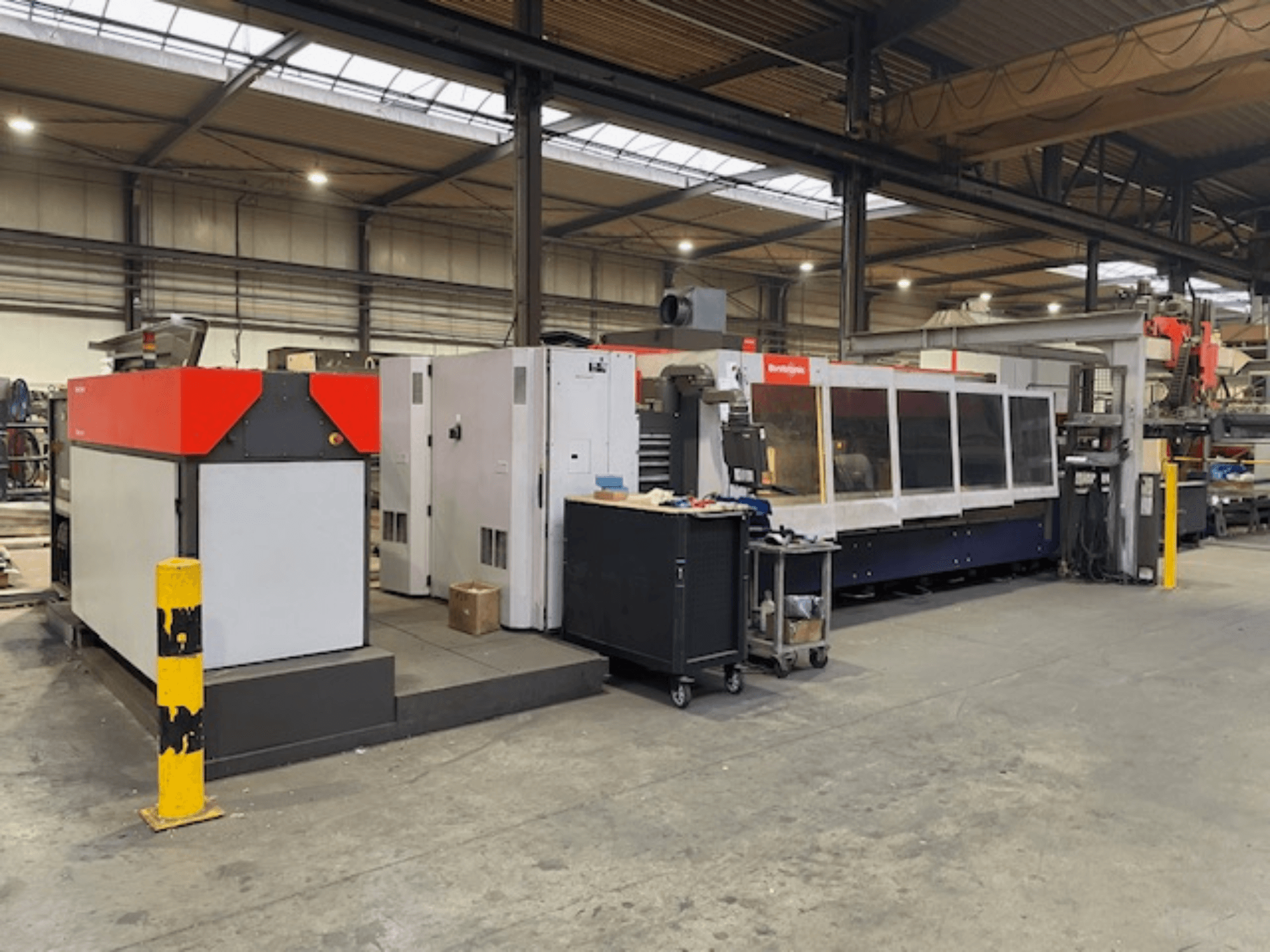 "Bystronic BySpeed 3015" CO2 lazerinio pjovimo staklės, šoninis vaizdas dirbtuvėse, su ryškiais raudonos ir pilkos spalvos dizaino elementais.