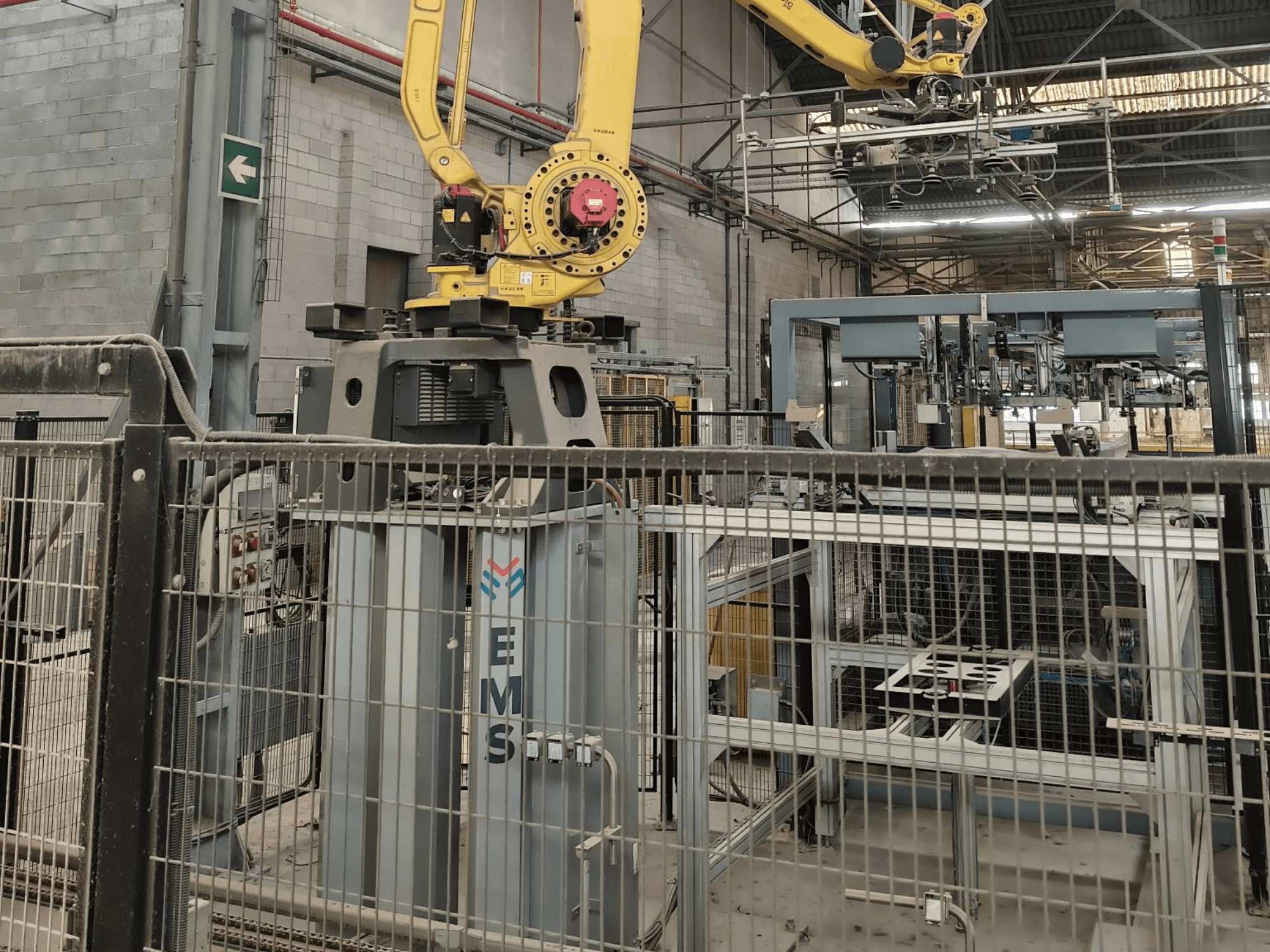 FANUC M-410iC/315 roboto rankos vaizdas iš priekio, kai ji yra virš EMS įrankio, o fone matoma pramoninė įranga ir aptvaras.