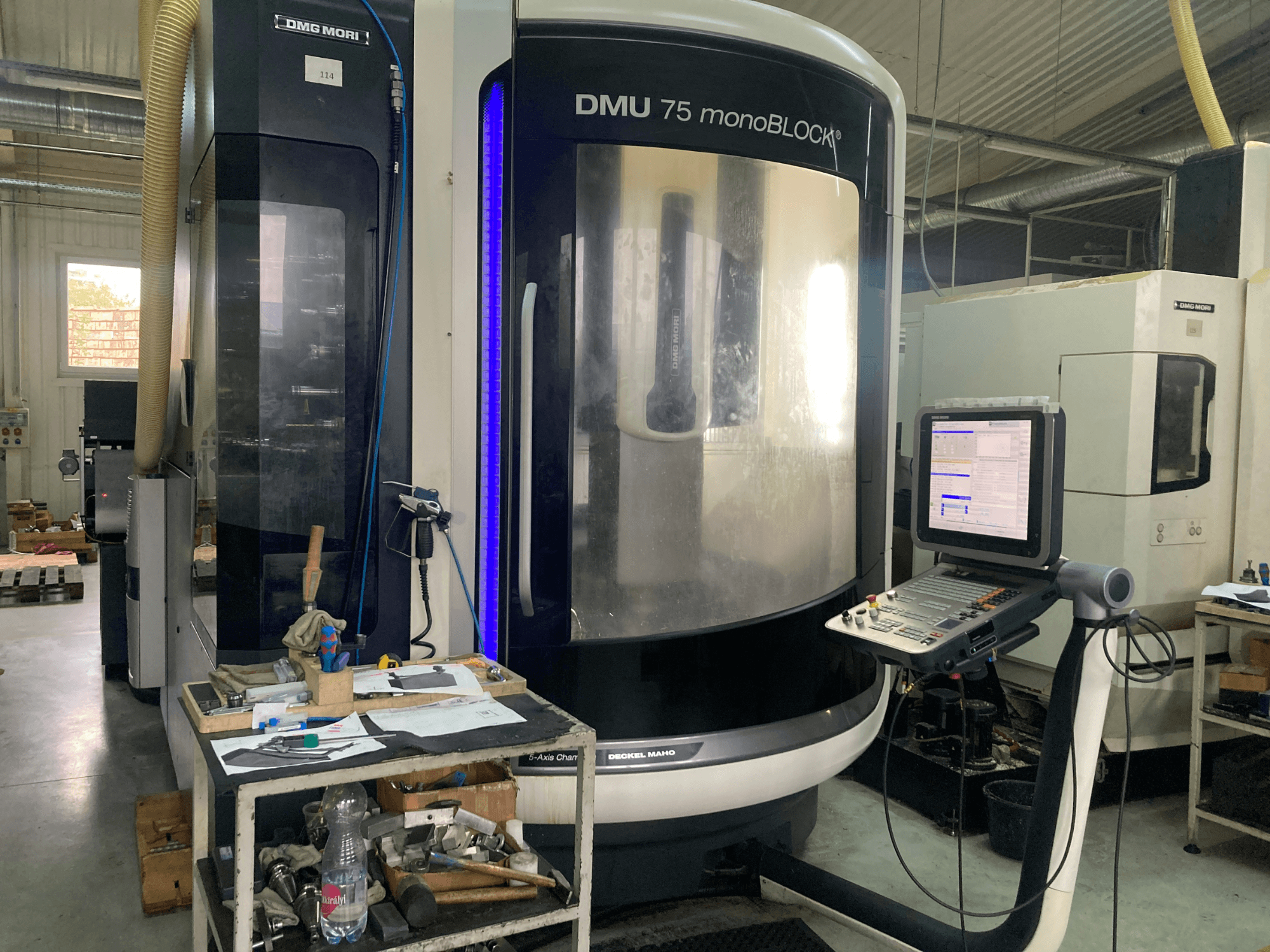 DMG MORI DMU 75 monoBLOCK® CNC staklės, vaizdas iš priekio: valdymo skydelis, įrankiai ir darbo vieta su medžiagomis.
