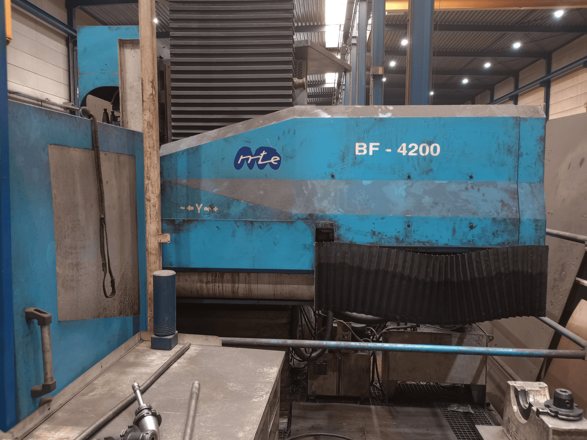 MTE BF 4200 lovinio tipo frezavimo staklės, vaizdas iš šono, kuriame matomas mėlynas korpusas ir išsamiai pavaizduotos staklių sudedamosios dalys dirbtuvės aplinkoje.