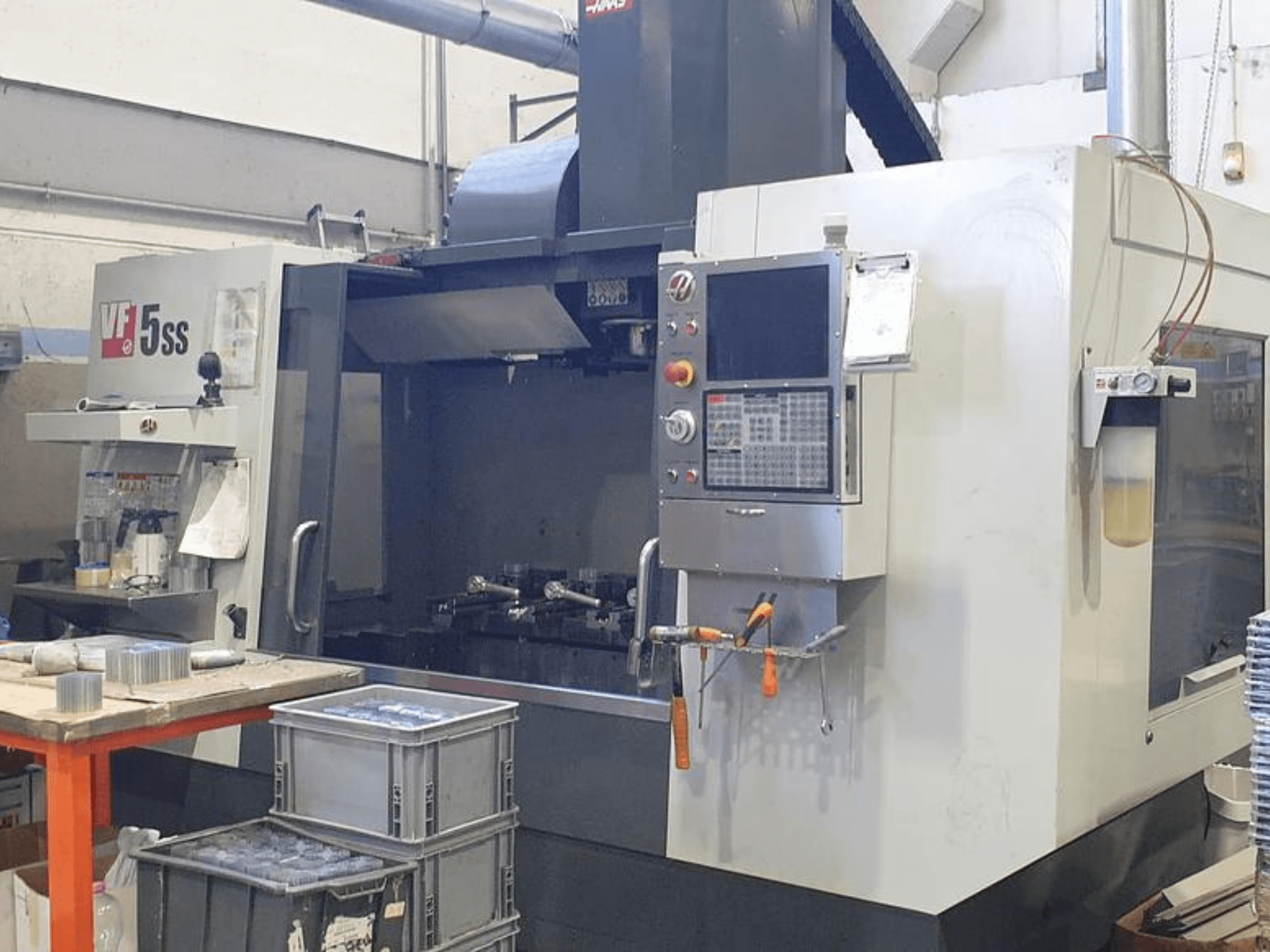 HAAS VF-5SS vertikalusis apdirbimo centras iš priekio, kuriame matyti valdymo skydelis, darbo vieta ir laikymo dėžės.