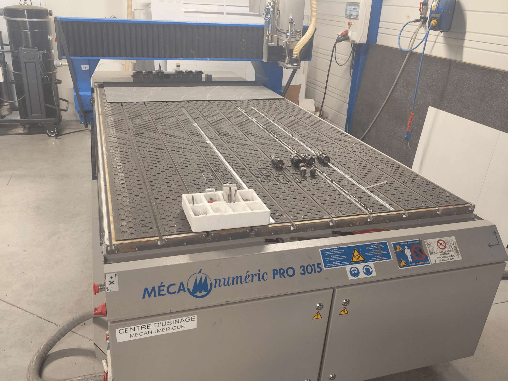 CNC frezavimo staklių „MECANUMERIC Pro 3015“ vaizdas iš priekio, kuriame matomas išsamiai pavaizduotas pjovimo stalas, valdymo pultas ir ant stalo esantys priedai.