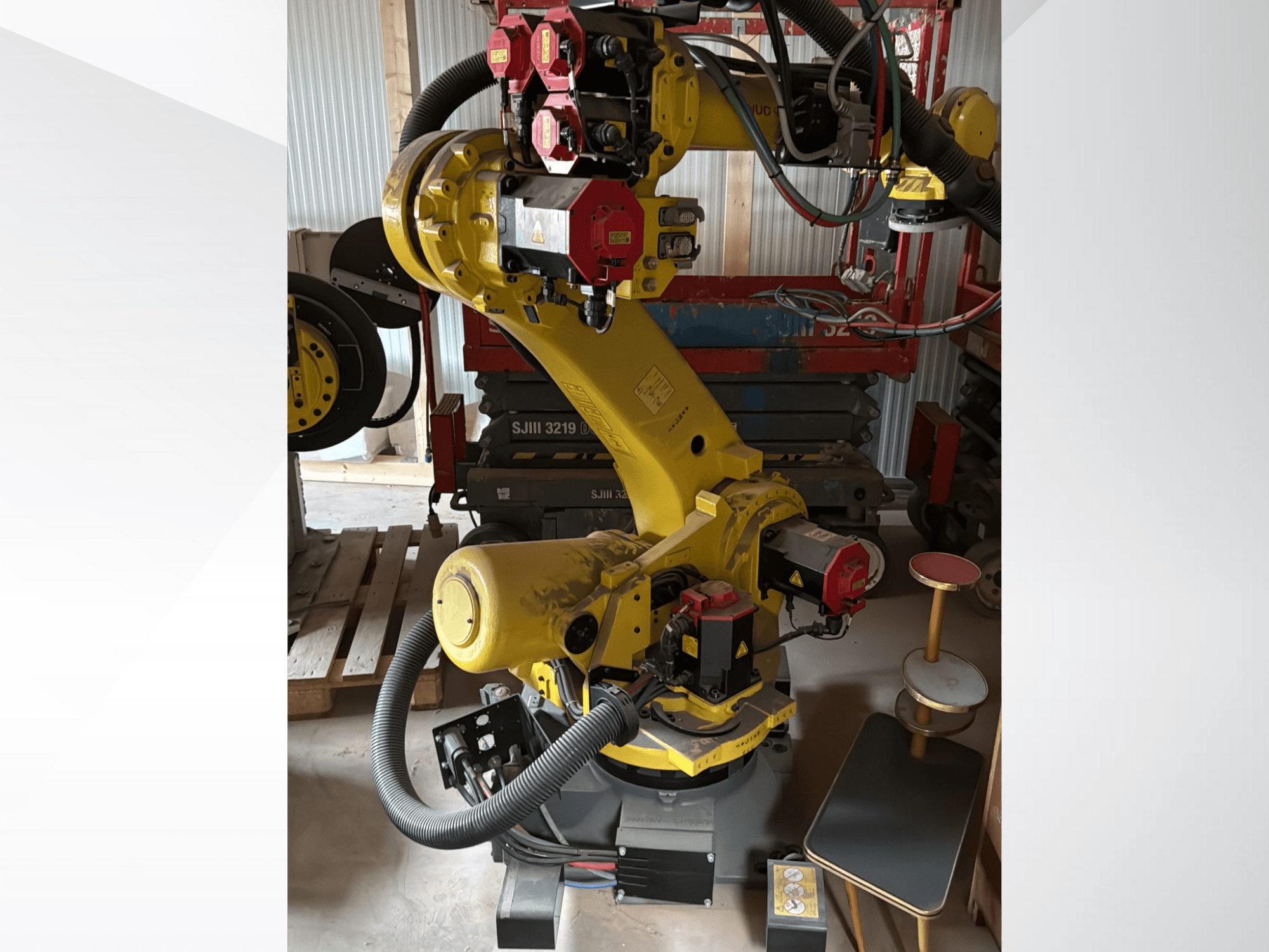 Geltonos pramoninės roboto rankos su raudonais valdikliais, vaizdas iš priekio; turi daugybę sąnarių ir žarnų, stovi darbo vietoje.