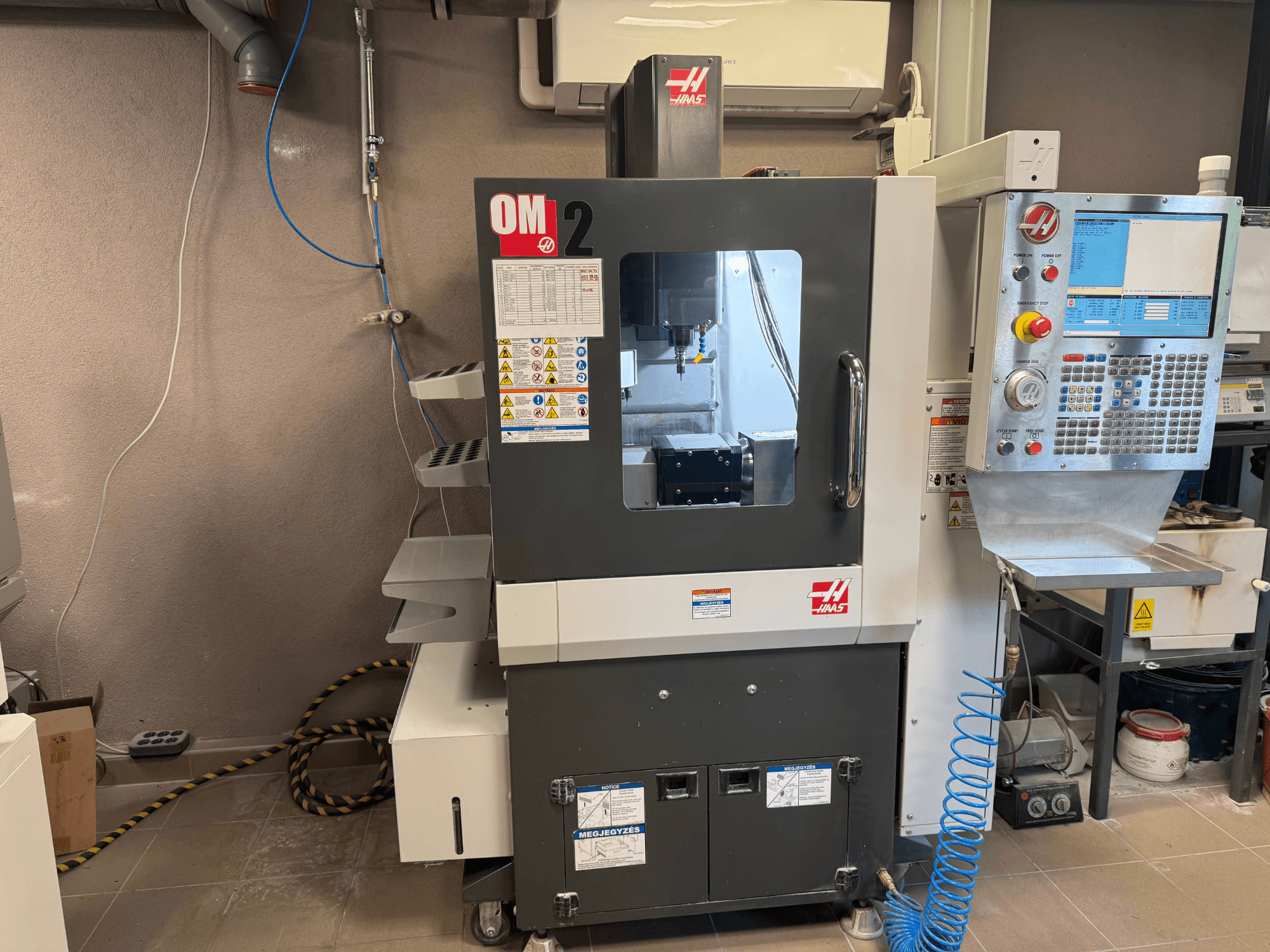 Haas OM-2 CNC staklės, vaizdas iš priekio, valdymo skydelis, suklys ir saugos etiketės, įrengtos dirbtuvių aplinkoje.