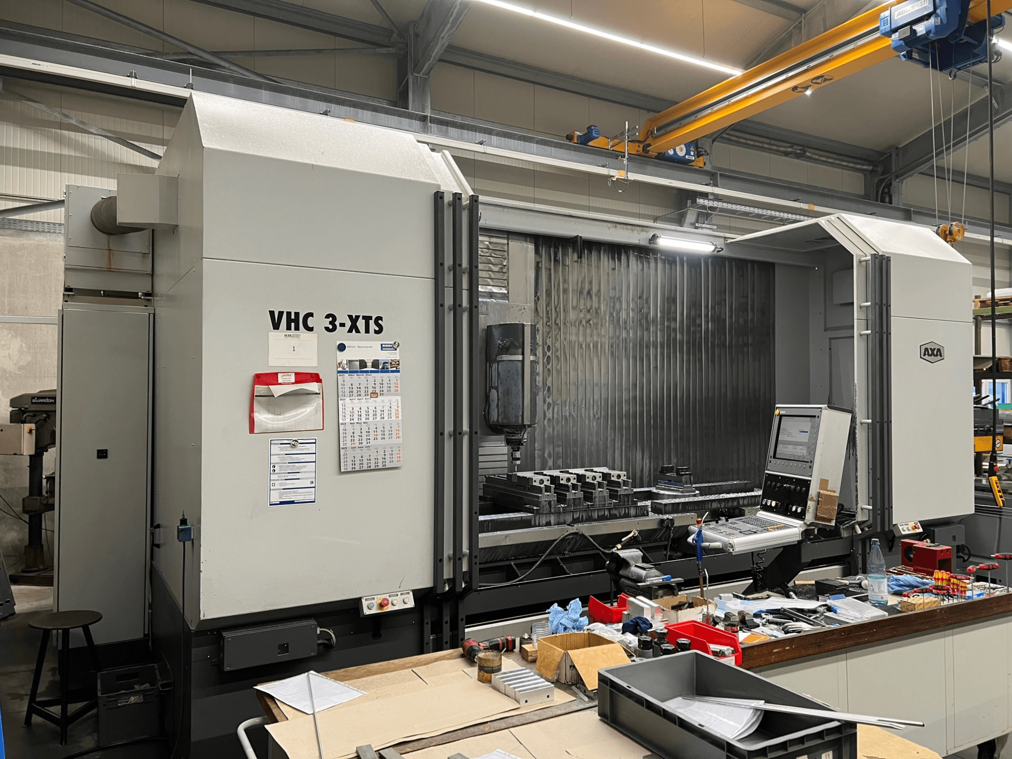 AXA VHC 3-XTS CNC staklės, žiūrint iš šono, su įrankių ir komponentų prigrūstu valdymo skydeliu ir darbo zona.