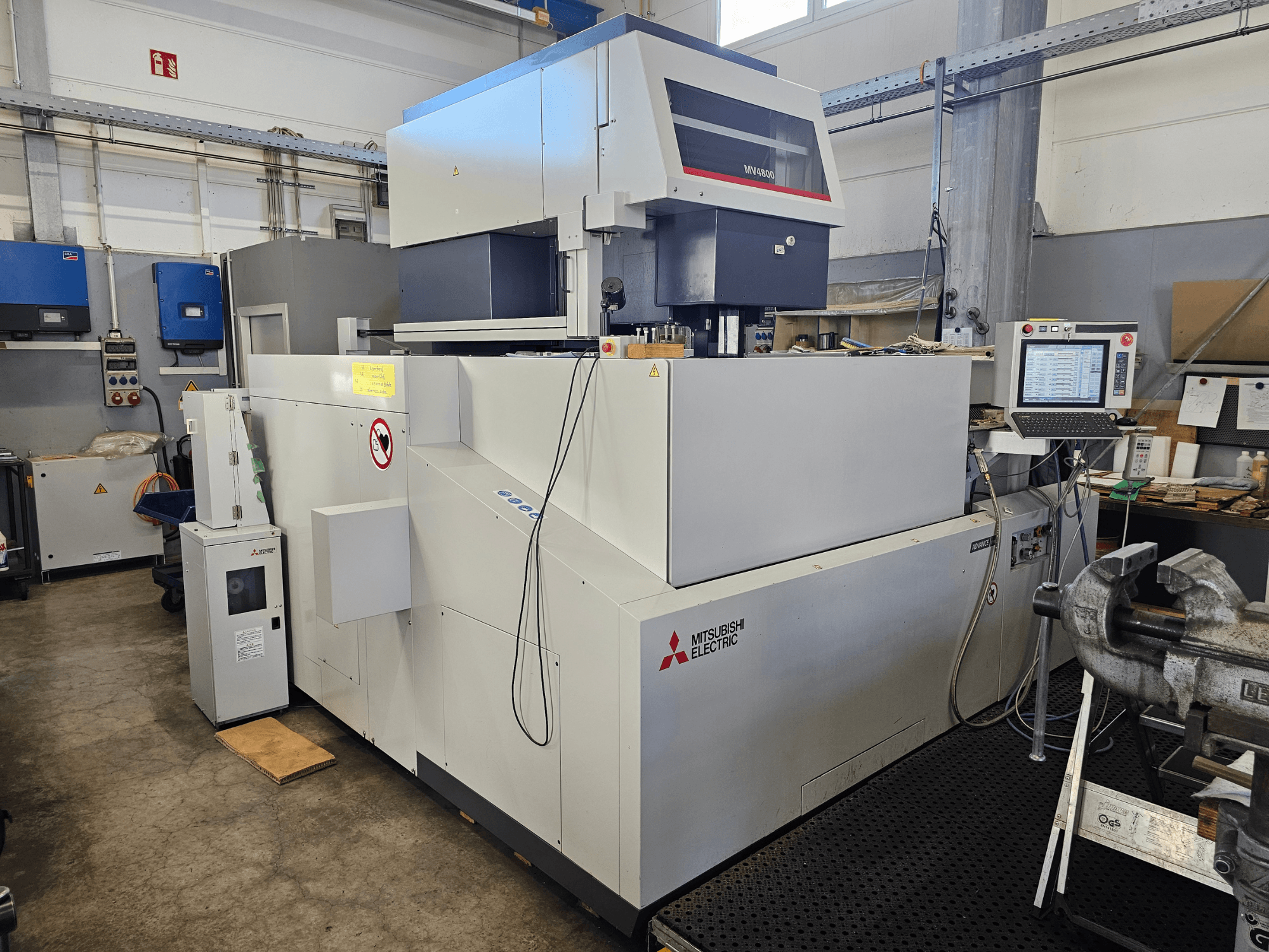 "Mitsubishi MV4800" vielos elektroerozinio apdirbimo staklės dirbtuvėse, šoninis vaizdas, kuriame matomas valdymo skydelis, saugos etiketės ir aplinkinė įranga.