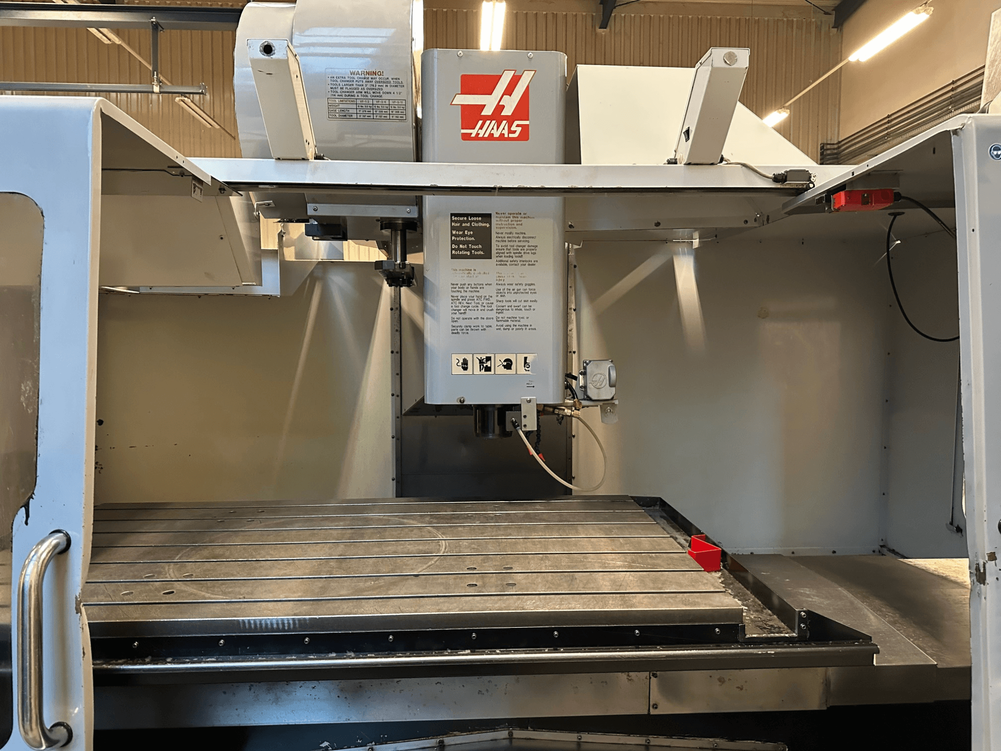 "Haas" CNC frezavimo staklės, modelis VF-2, priekinis vaizdas, kuriame matomi valdymo įtaisai, metalinis darbo stalas ir saugos etiketė ant skydelio.