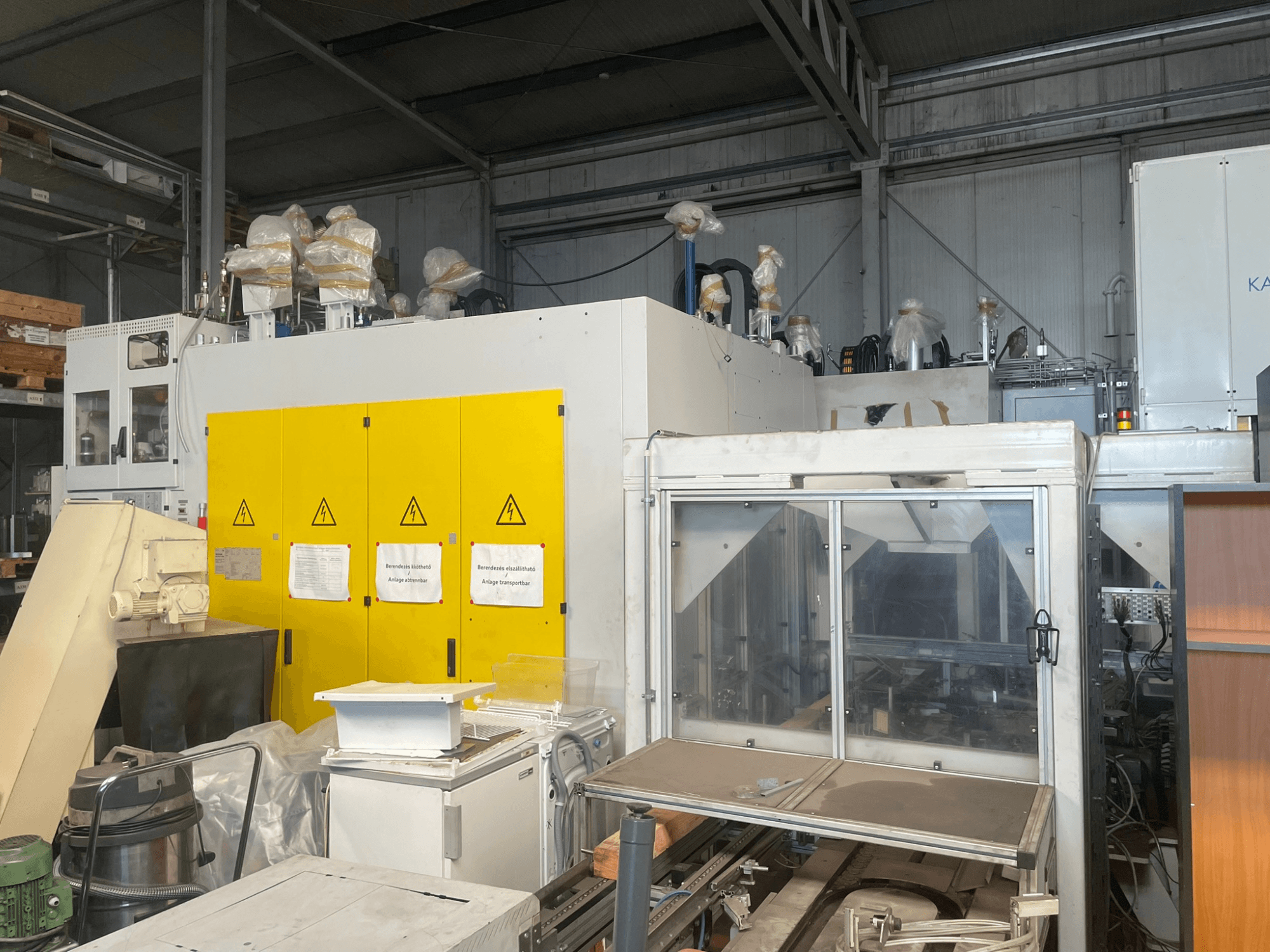 Pramoninės mašinos su geltonu valdymo skydeliu, vaizdas iš šono, su įspėjamaisiais užrašais ir plastikinėmis dangomis, sandėlyje.