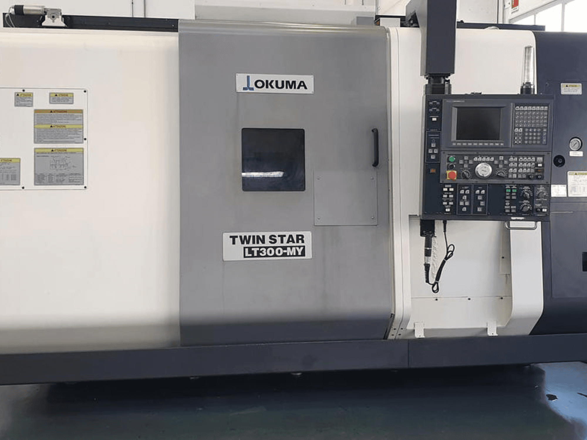 "Okuma Twin Star LT300-MY" CNC tekinimo staklės iš šono, su valdymo skydeliu, valdymo mygtukais ir saugos etiketėmis.