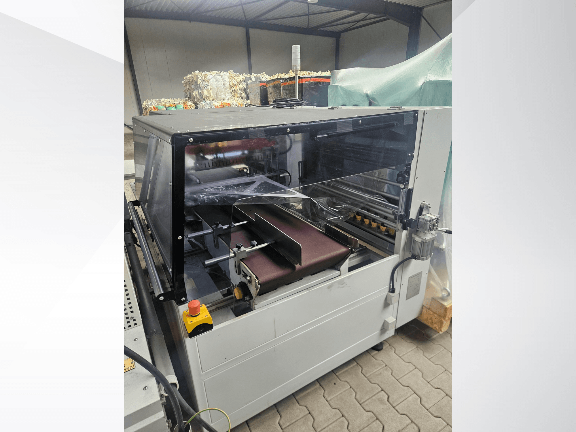 „AMTEC Speed Shrink 40 PRO“ mašina, vaizdas iš priekio, kuriame matomas plastiko apdorojimo mechanizmas su skaidriu apsauginiu gaubtu ir konvejeriu.