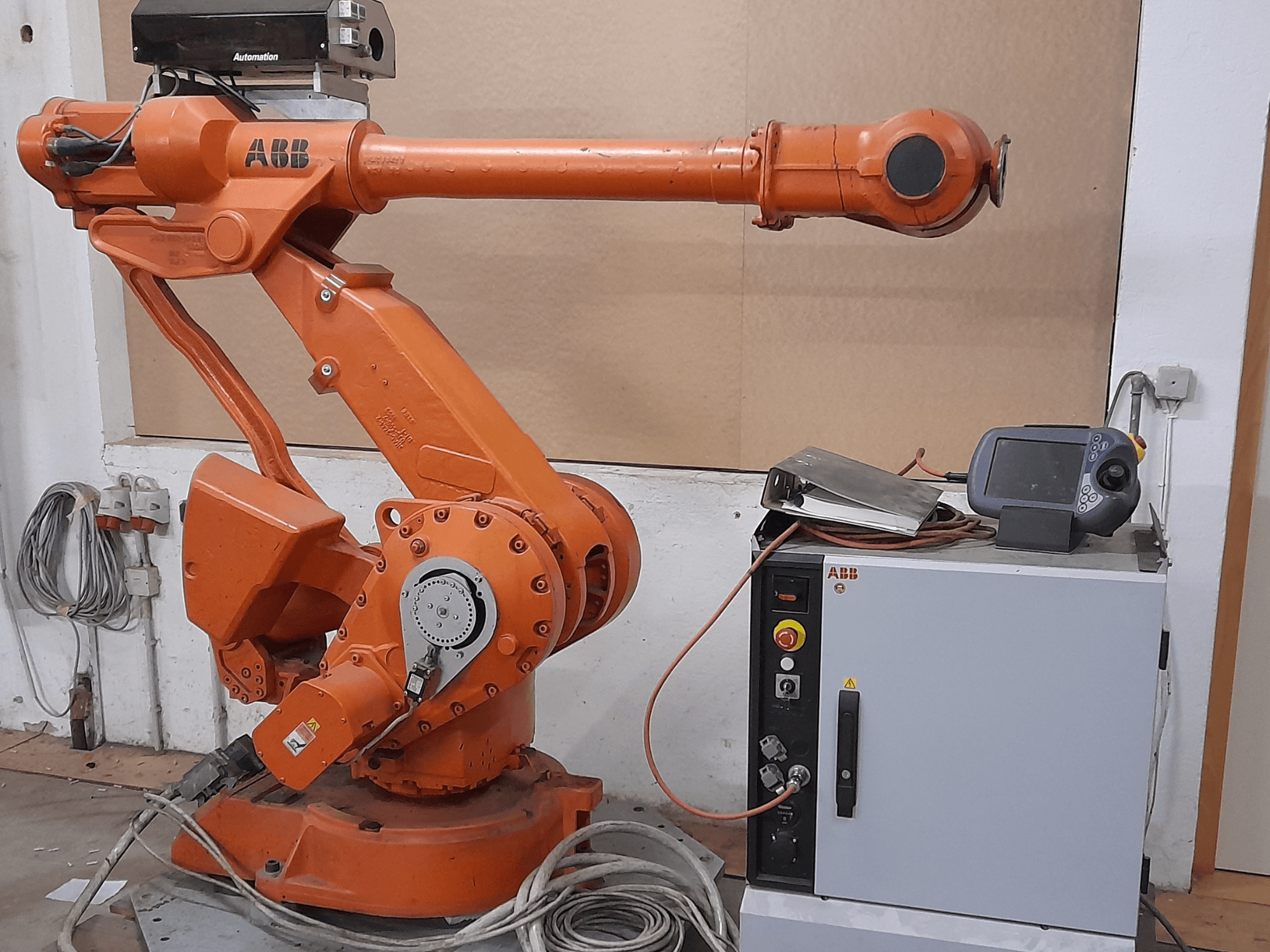 Oranžinė ABB roboto ranka su valdymo skydeliu, vaizdas iš priekio, demonstruojant kabelius ir automatikos įrangą.